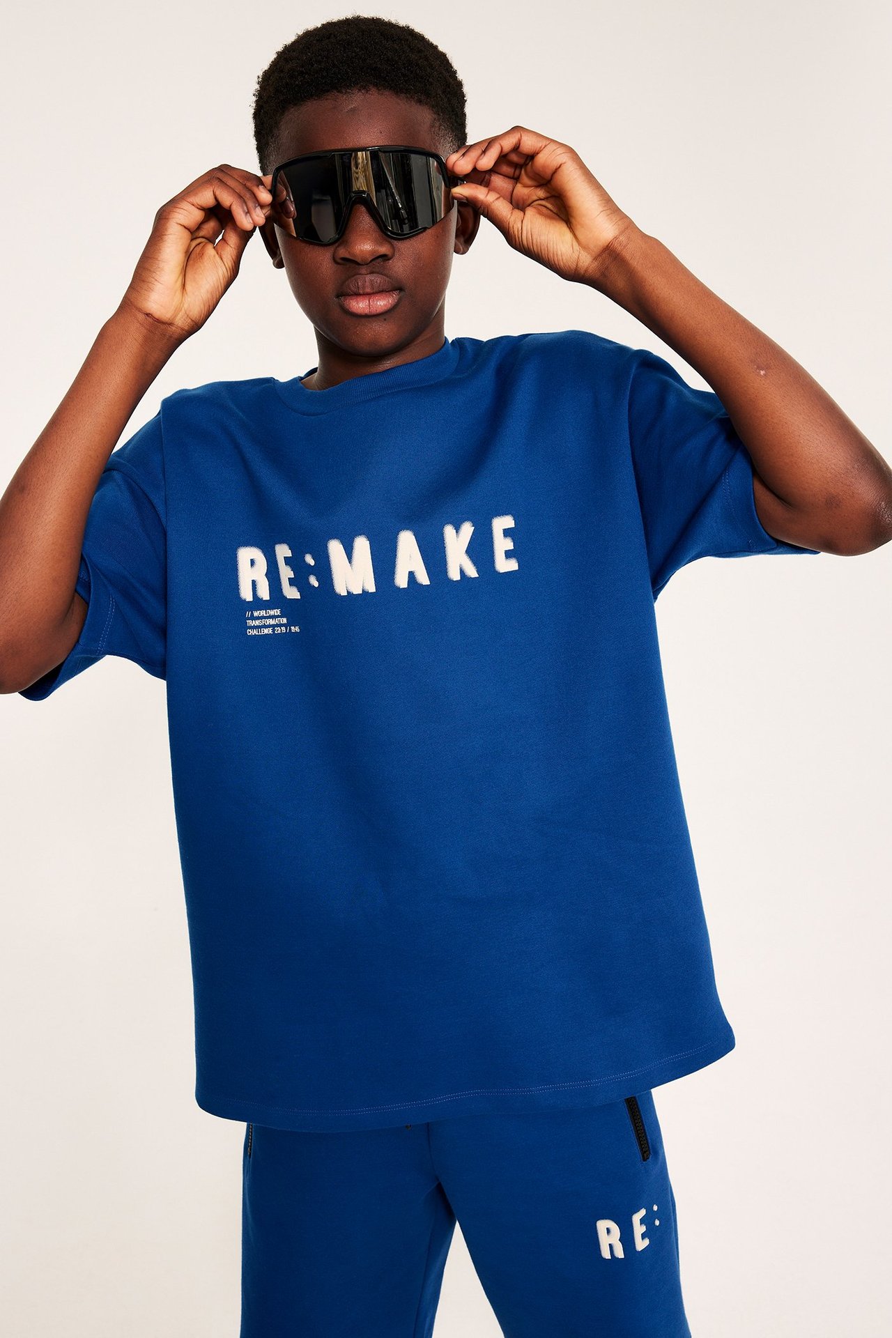 Widok z przodu: Dziecko w granatowym, oversize’owym t-shircie „RE:MAKE” i pasujących dresach, poprawiające czarne okulary.