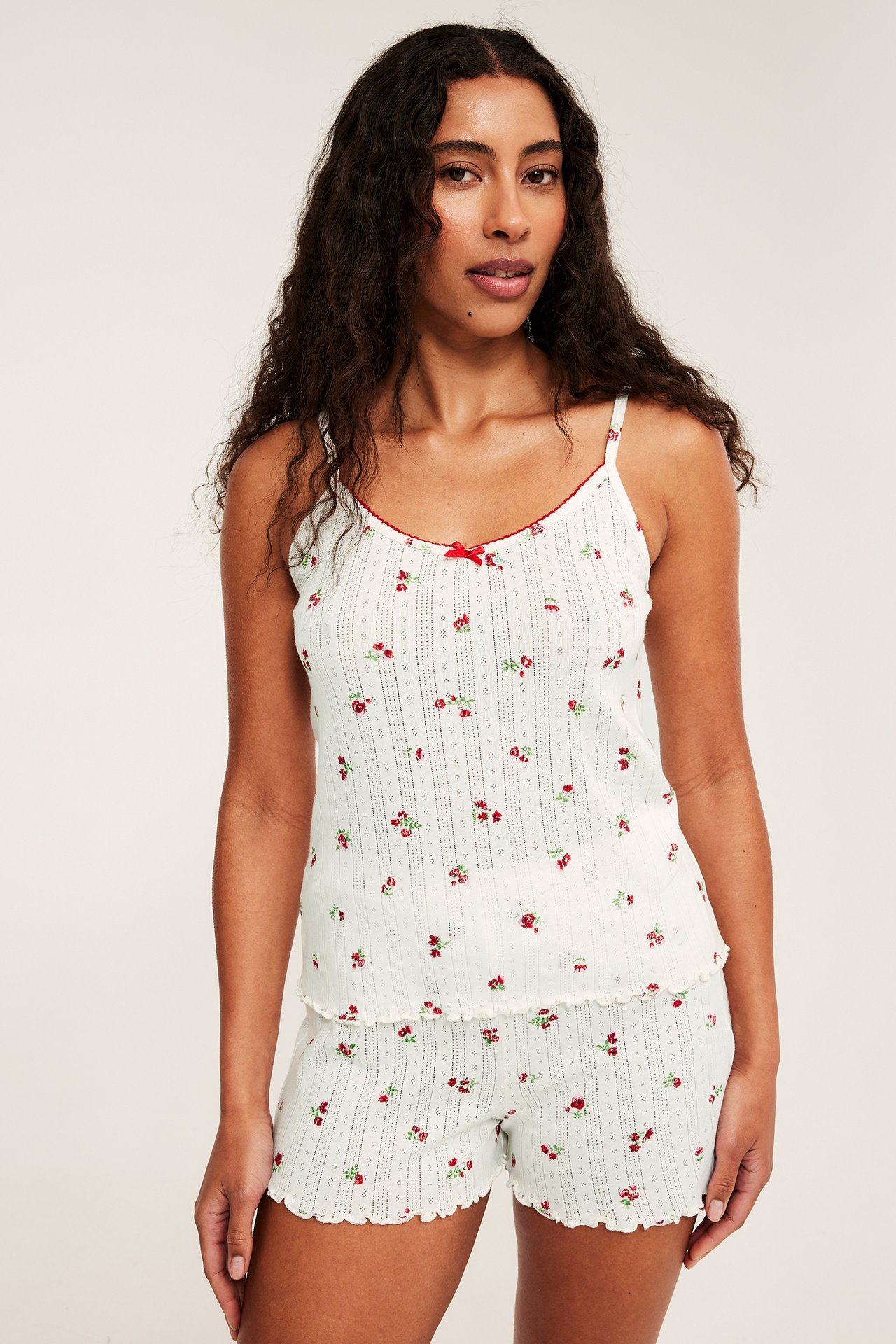 Forfra: kvinne i hvit bomulls-pointelle pyjamas med rødt blomstertrykk, topp med spaghetti-stropper og shorts.