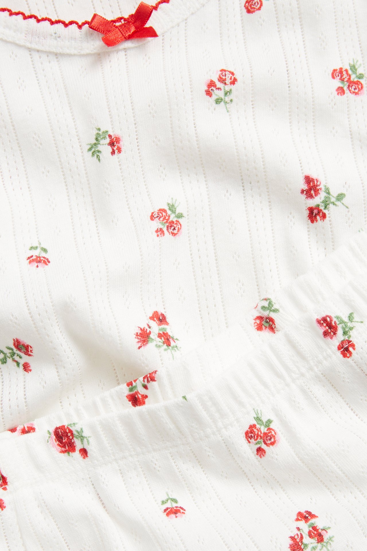 Nærbilde av hvit bomulls-pointelle pysjamasett med rødt blomstertrykk, rød kant og liten sløyfe.