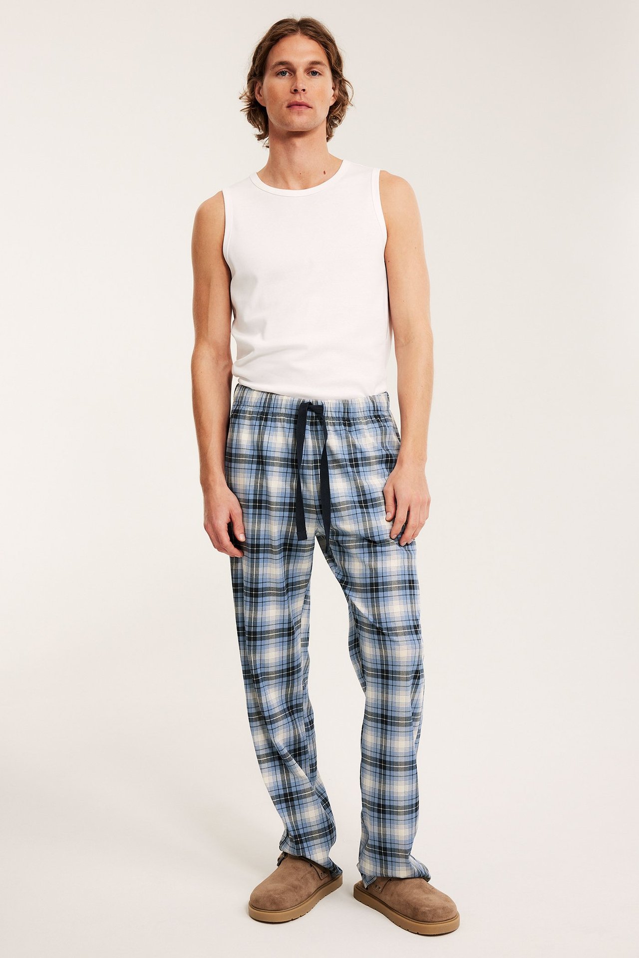 Frontvy av en man i blårutiga pyjamasbyxor i bomull, vitt linne och bruna tofflor.