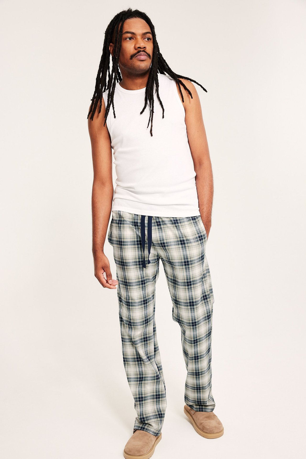 Mężczyzna przodem w ciemnozielonych, kraciastych spodniach piżamowych z bawełny, białym topie i brązowych kapciach.