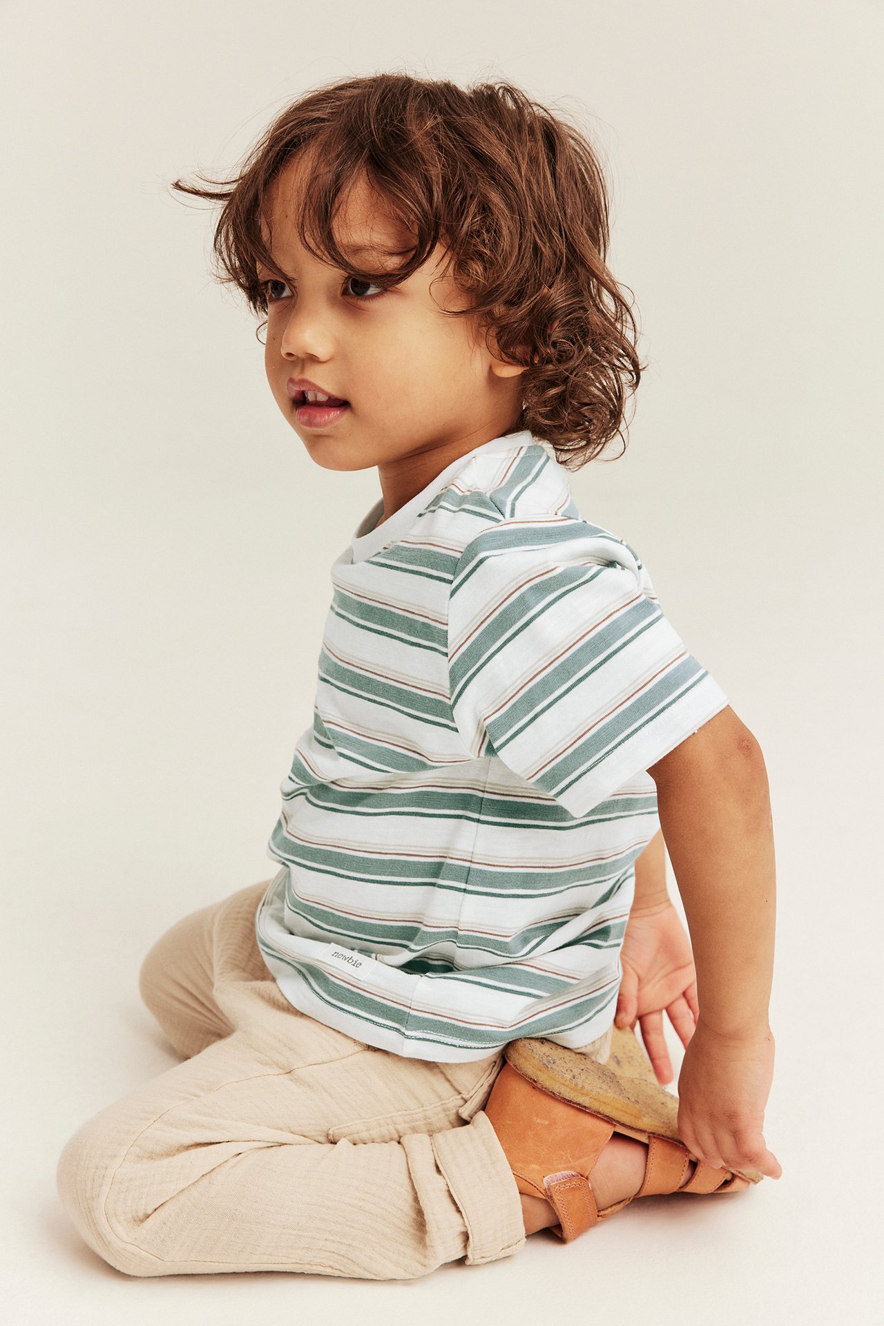 Sidovy av ett barn i randig kortärmad offwhite slubtrikå t-shirt, beige byxor och bruna sandaler.