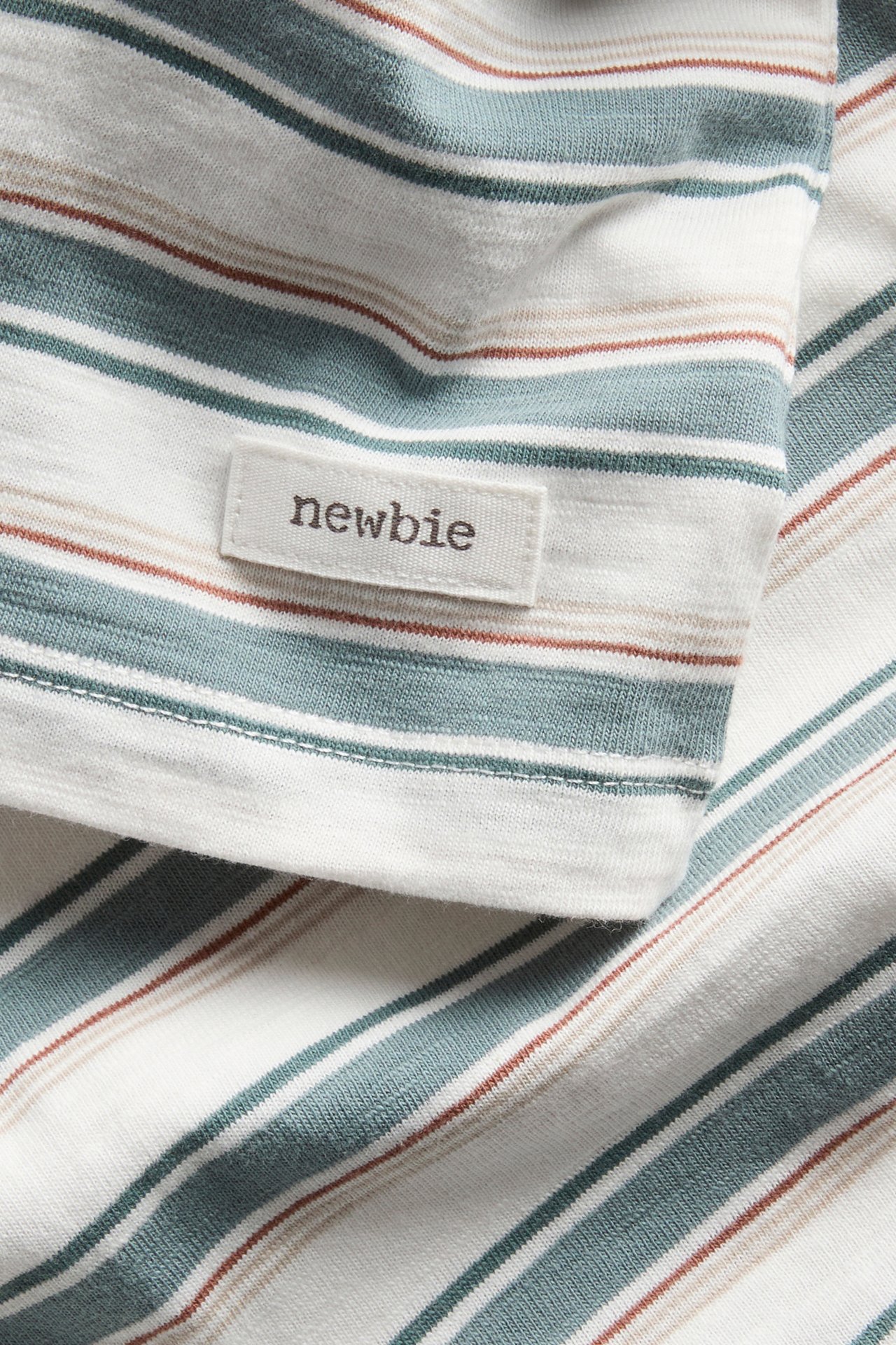 Zbliżenie na biało-kremowy t-shirt ze splotu slub w paski turkusowe, brązowe i beżowe, metka Newbie.