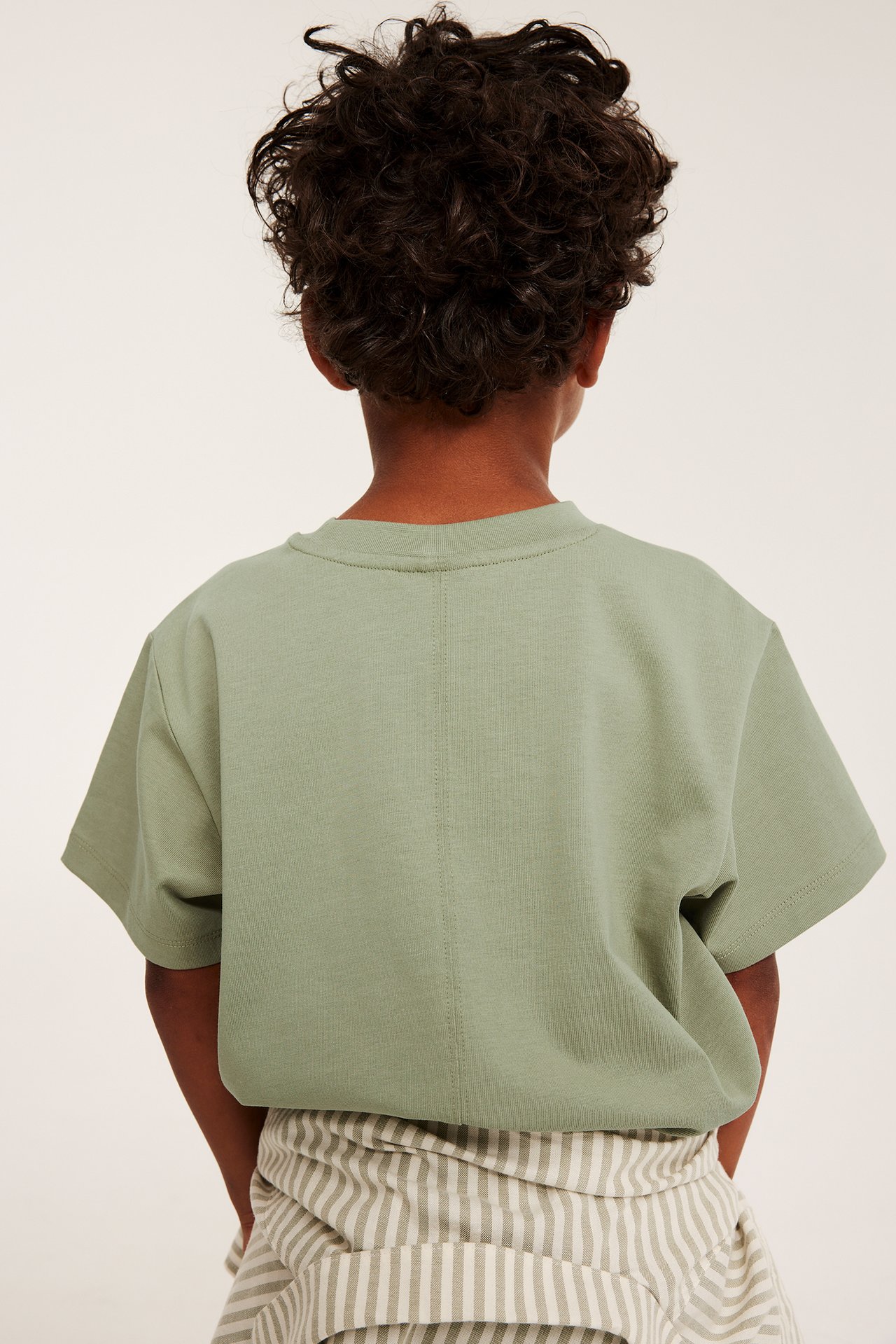 Lapsen takakuva tummanvihreässä oversized puuvilla-T-paidassa ja beige-vihreäraidallisissa housuissa.