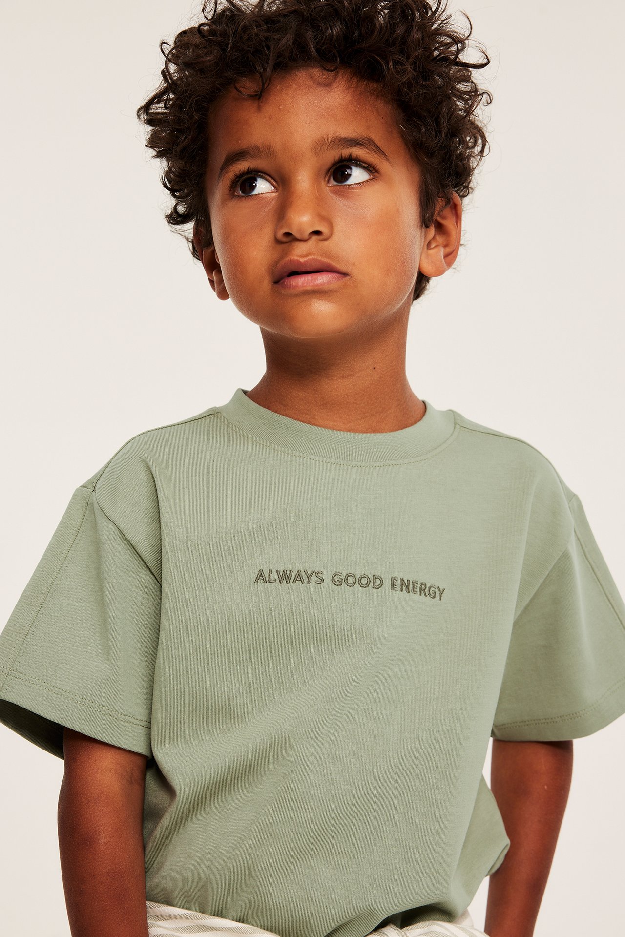 Framifrån: Kid bär mörkgrön oversized t-shirt i bomull med broderad text 'ALWAYS GOOD ENERGY'.