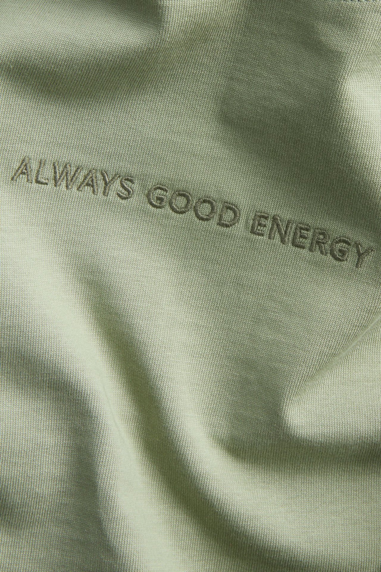 Närbild på en mörkgrön oversized t-shirt i bomull med broderad text "ALWAYS GOOD ENERGY".