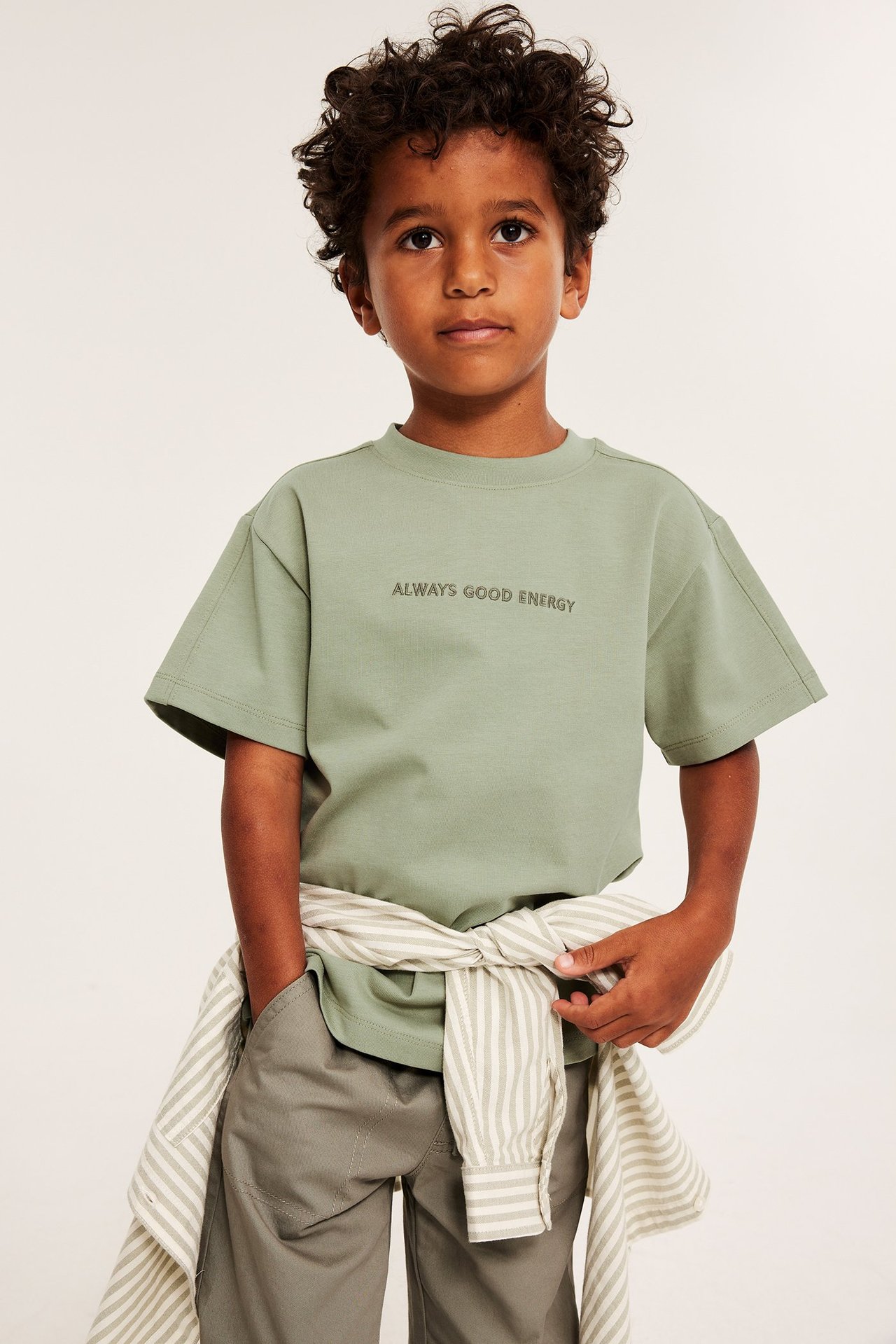 Ett barn i mörkgrön oversized t-shirt med broderi, grå byxor och randig skjorta knuten runt midjan.