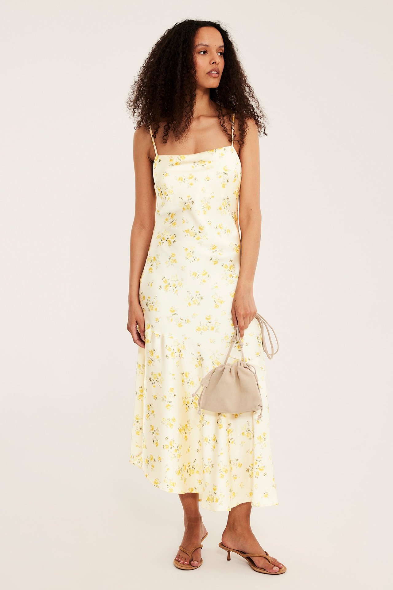 Framsida: kvinna i ljusgul blommig jacquard slipklänning, håller beige dragskoväska, bär bruna klacksandaler.