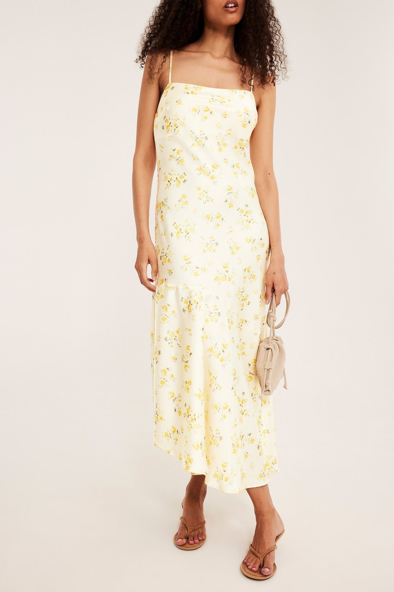 Frontvy av kvinna i ljusgul blommig jacquard slipklänning, håller beige handväska, bär bruna flip-flops.