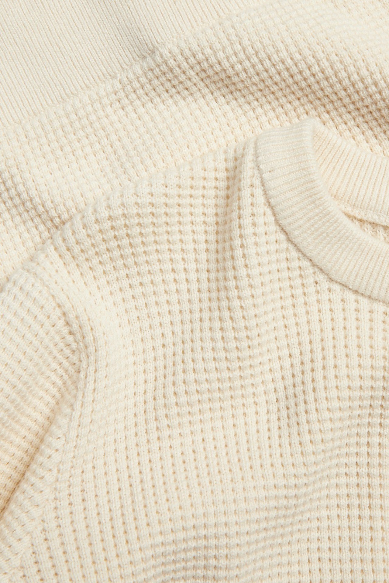 Närbild av en ljusbeige finstickad långärmad tröja i bomull med våffelmönster och ribbad halsringning.