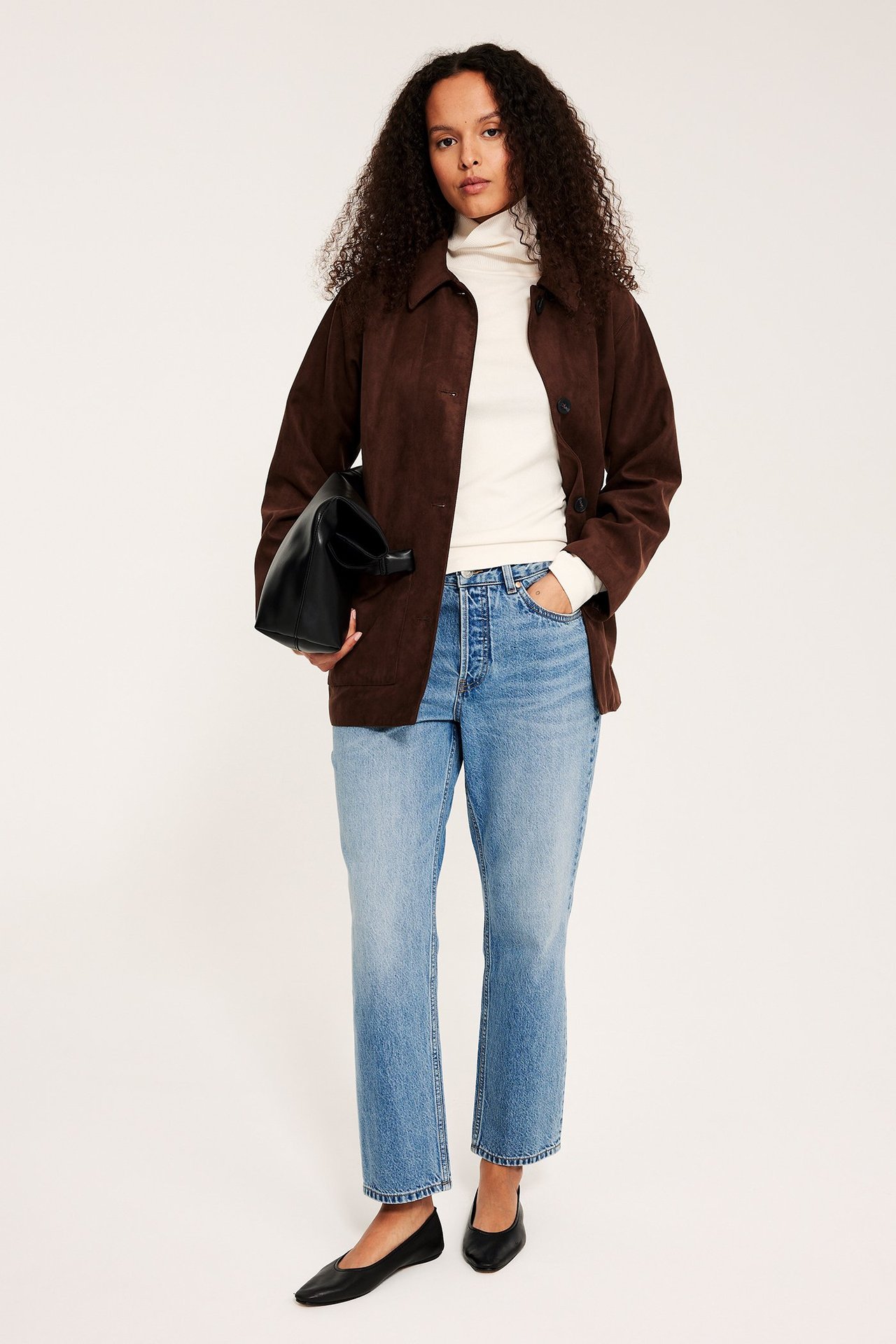 Kobieta w jasnoniebieskich prostych dżinsach z wysokim stanem, brązowej kurtce, kremowym golfie, czarnej torbie, czarnych baletkach.