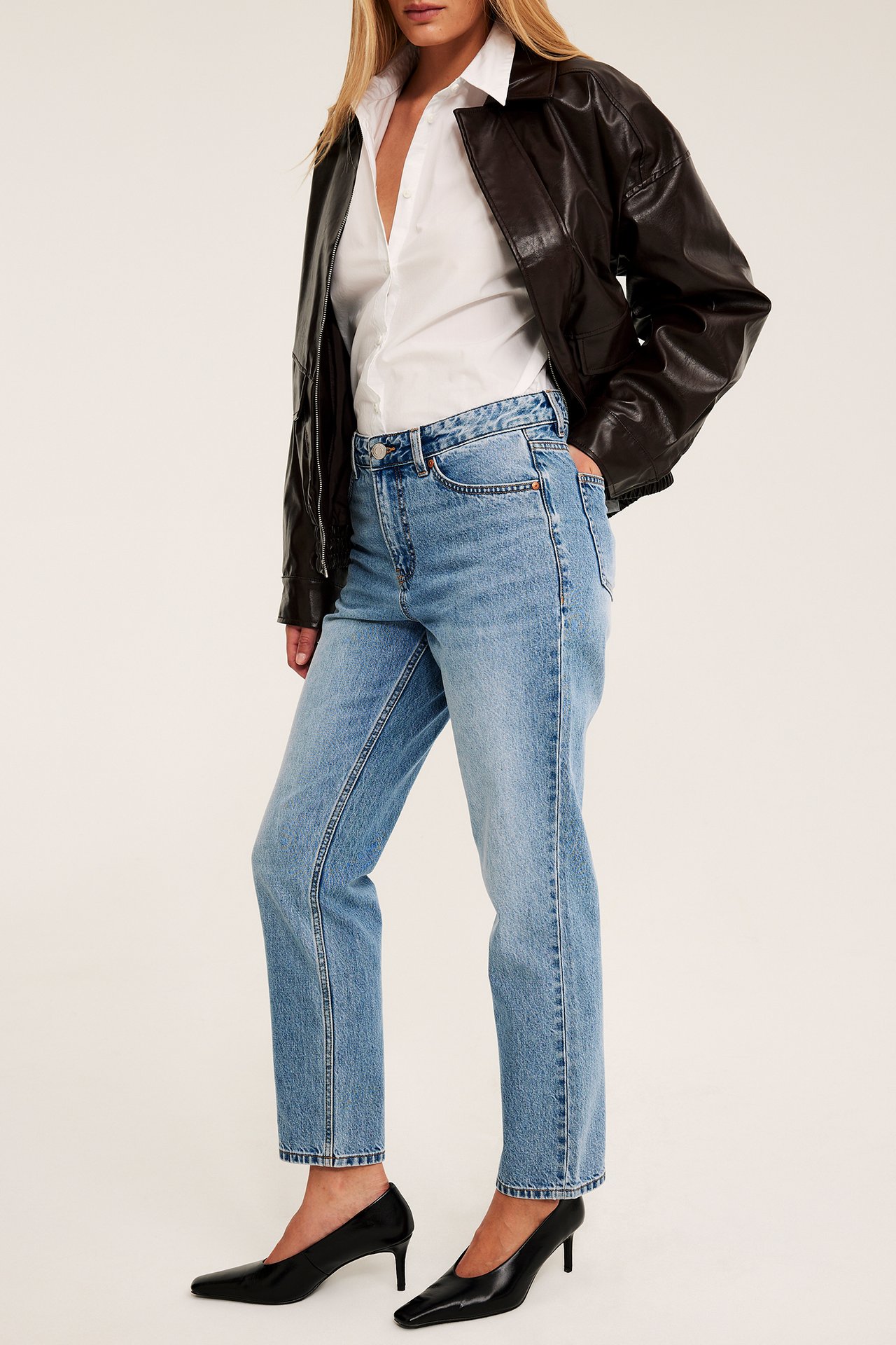 Kvinne i lyseblå rette jeans, hvit skjorte, brun skinnjakke og svarte hæler.