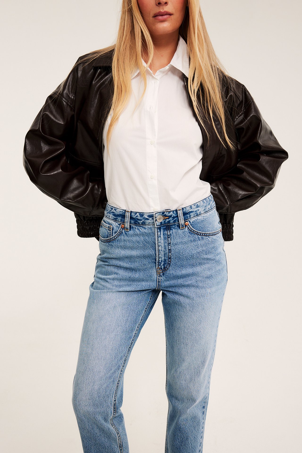 Forfra, kvinne i svart bomberjakke i kunstskinn, hvit skjorte og lyseblå jeans.