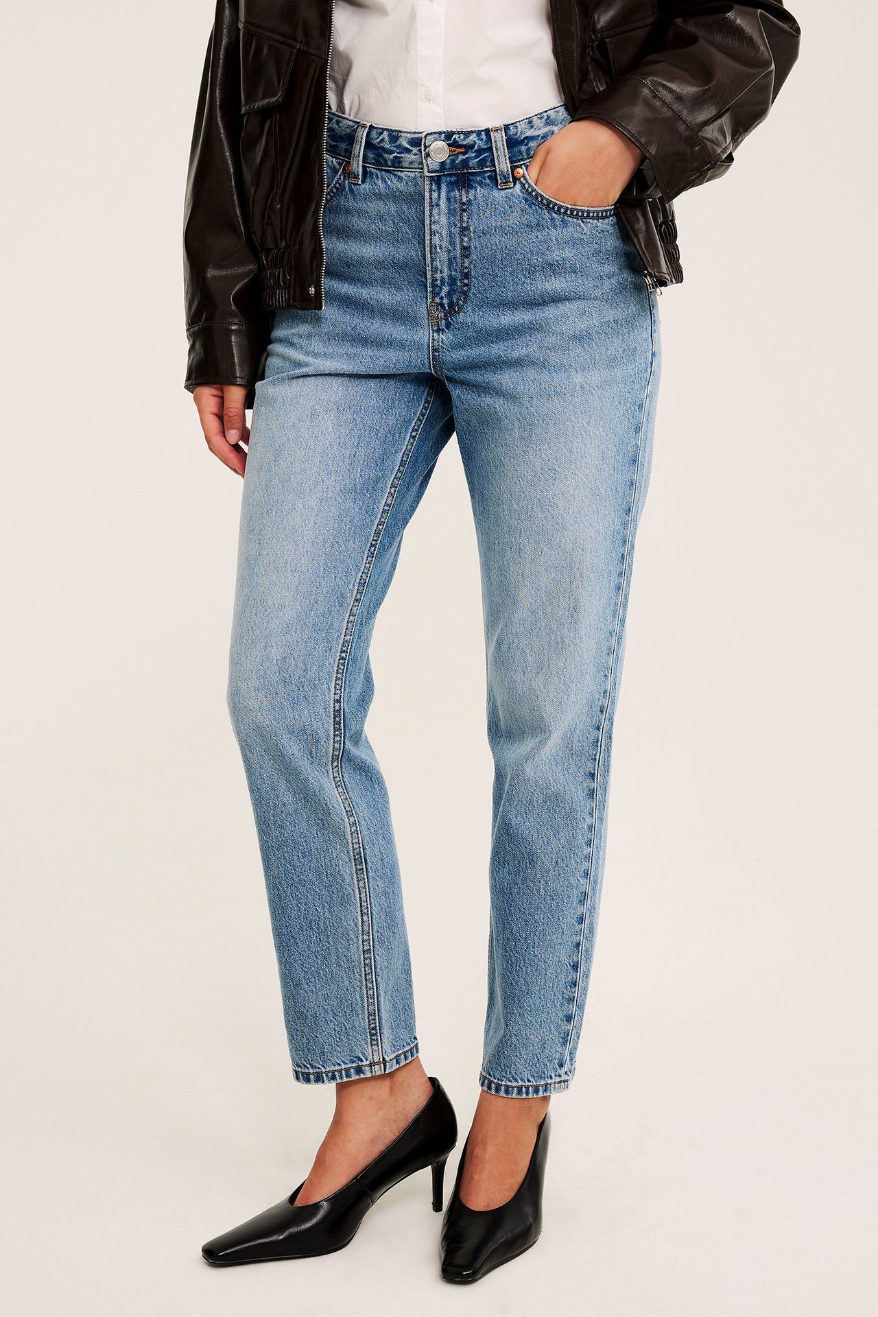 Front av kvinne iført lys blå rette jeans, svart skinnjakke, hvit skjorte og svarte hæler.