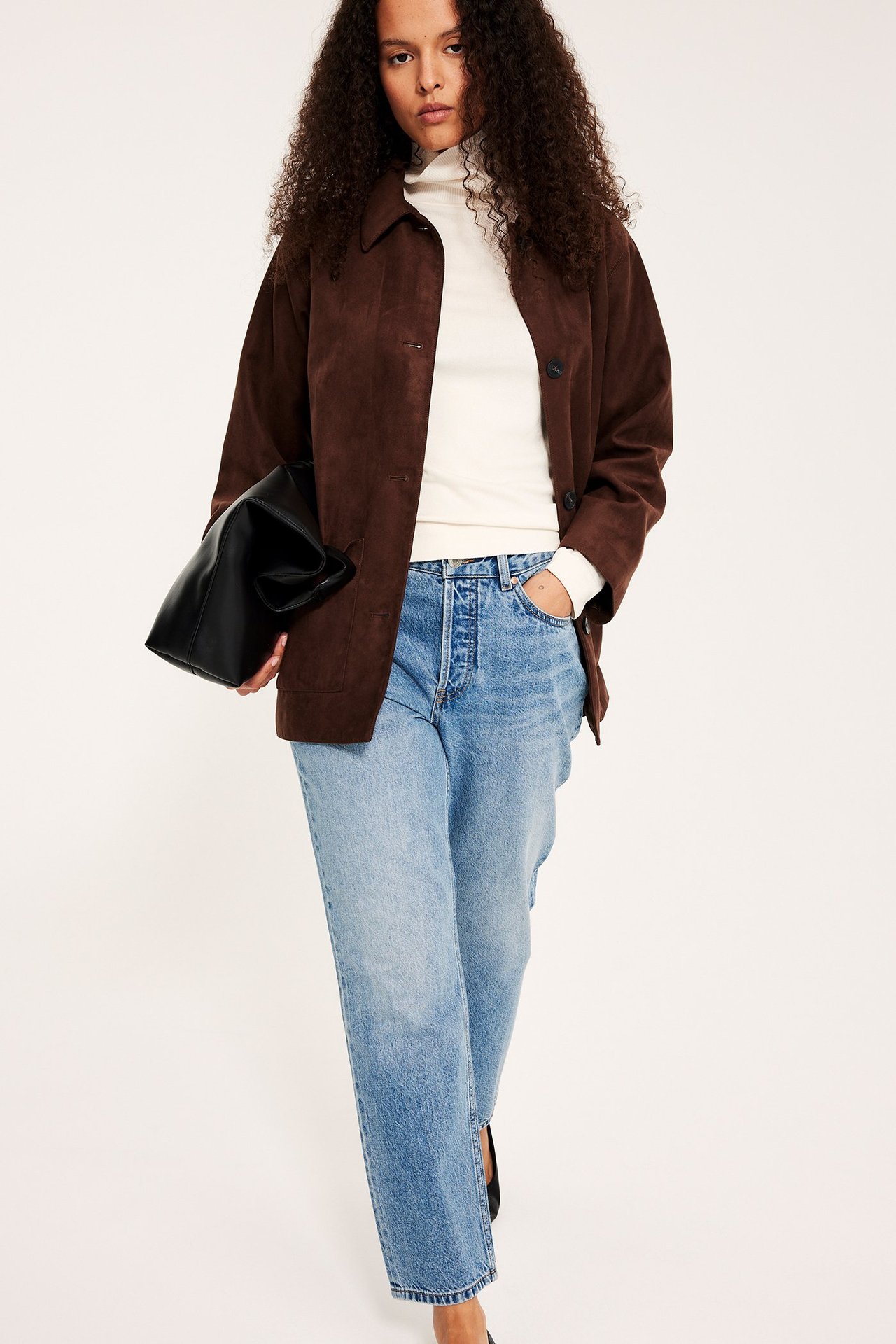 Kobieta ma na sobie jasnoniebieskie, proste, z wysokim stanem dżinsy cropped, brązową kurtkę, biały golf i czarną torebkę.