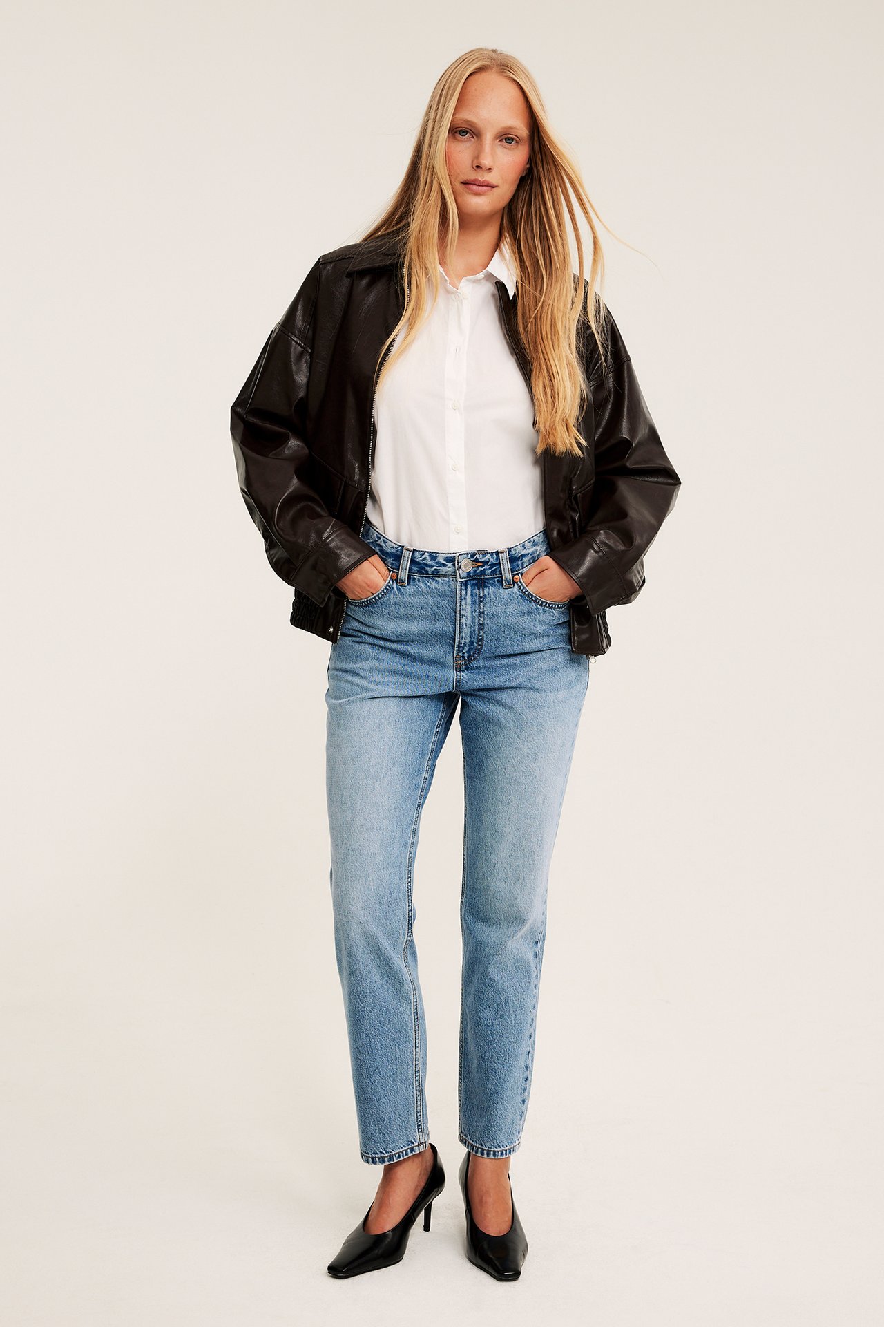 Frontvy av kvinnlig modell i mörkbrun skinnjacka, vit skjorta, blå jeans och svarta klackskor.