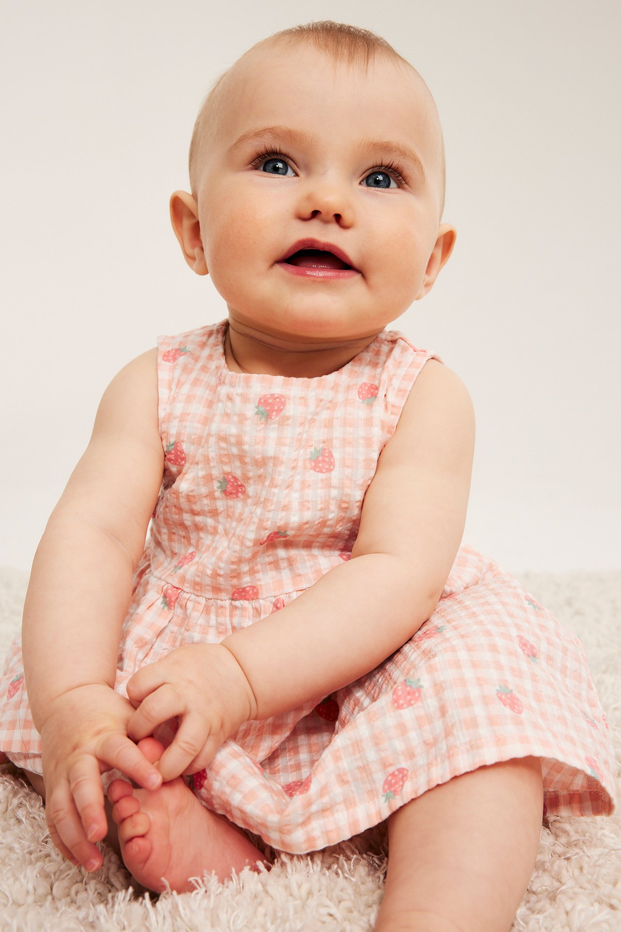 Forfra, baby i lyserosa seersucker-kjole med hvit gingham- og jordbærmønster.