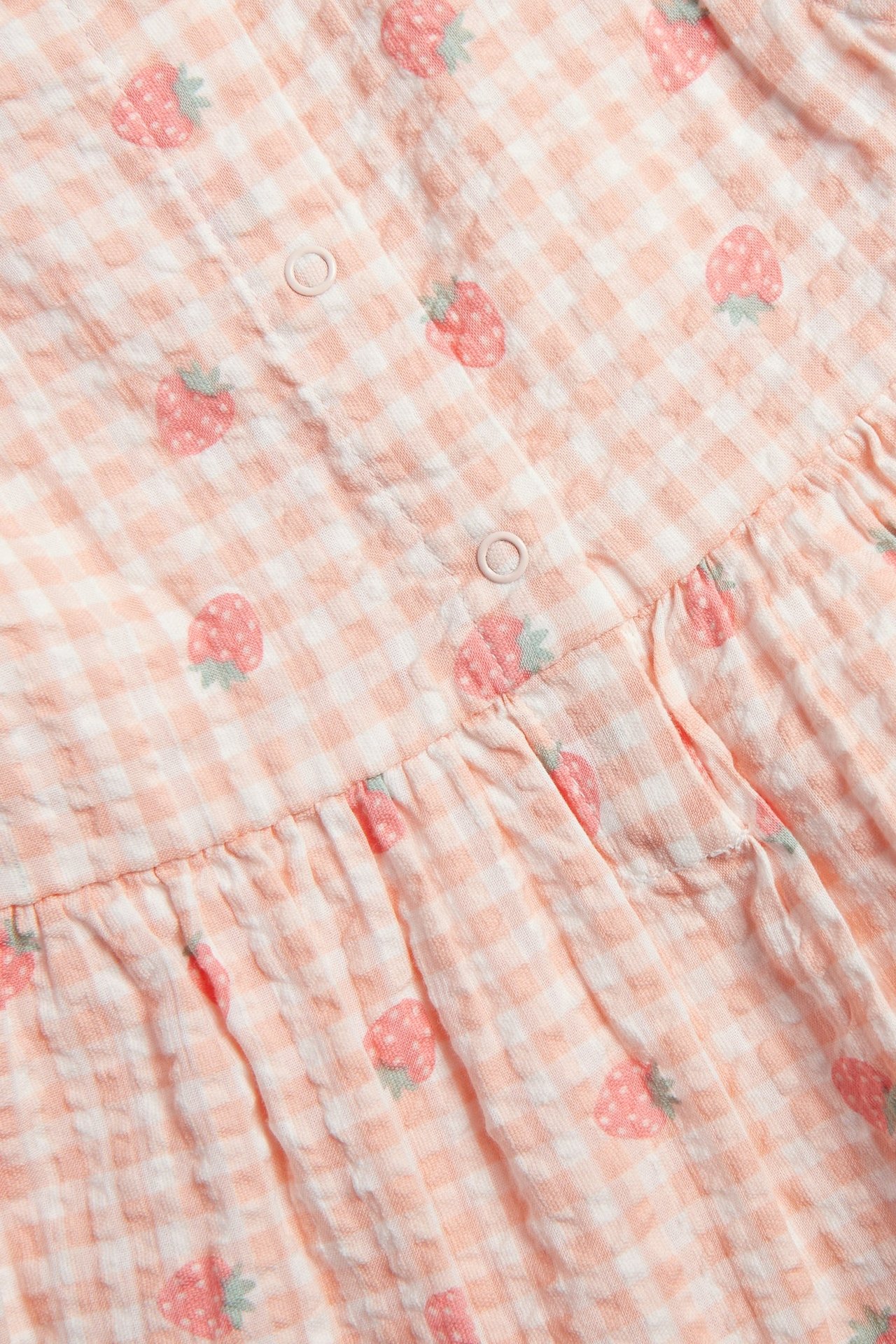 Zbliżenie jasno-różowej sukienki seersucker dla niemowląt w kratkę z wzorem truskawek i zatrzaskami.