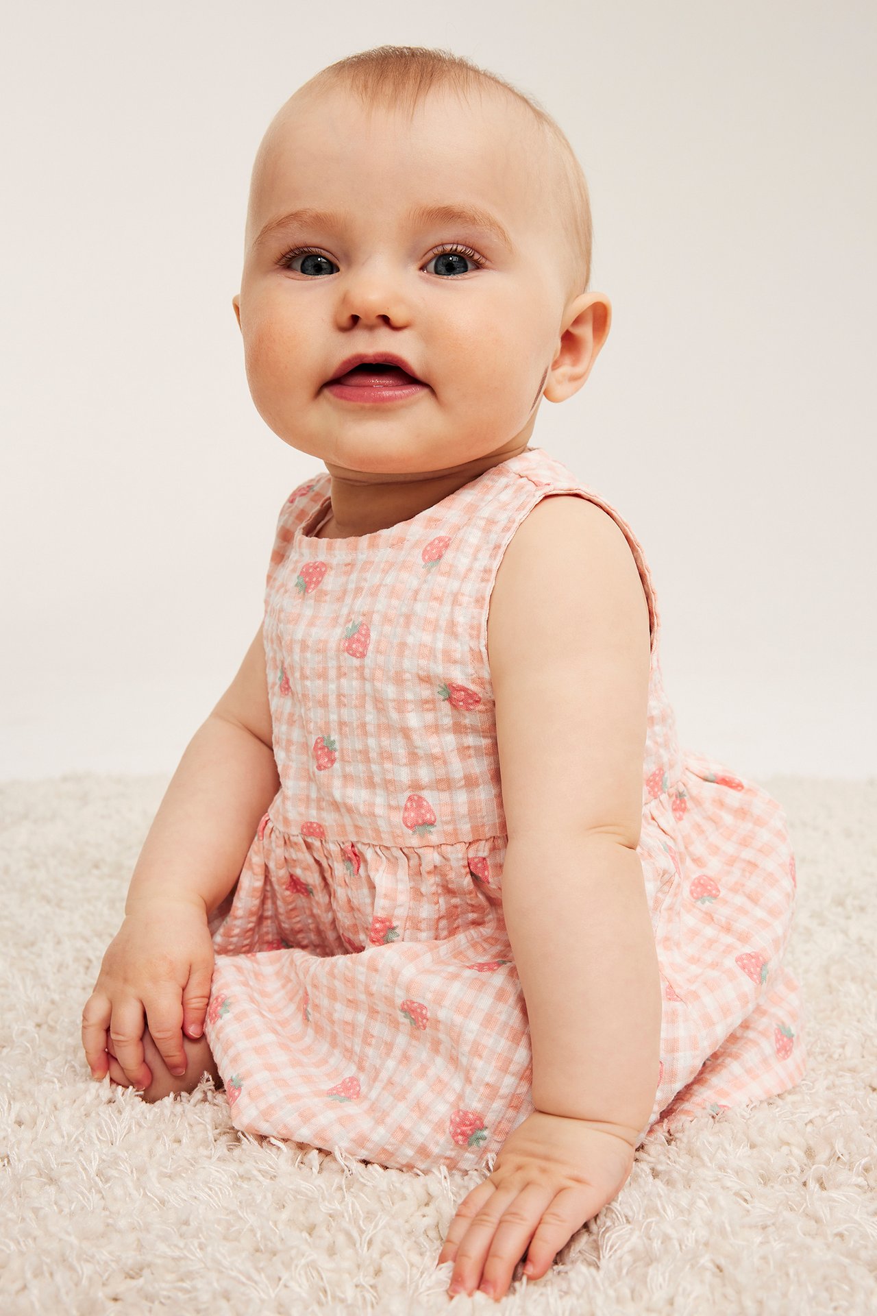 Widok z przodu: siedzące niemowlę w jasnoróżowej, kratkowanej bawełnianej sukience seersucker z truskawkowym wzorem.