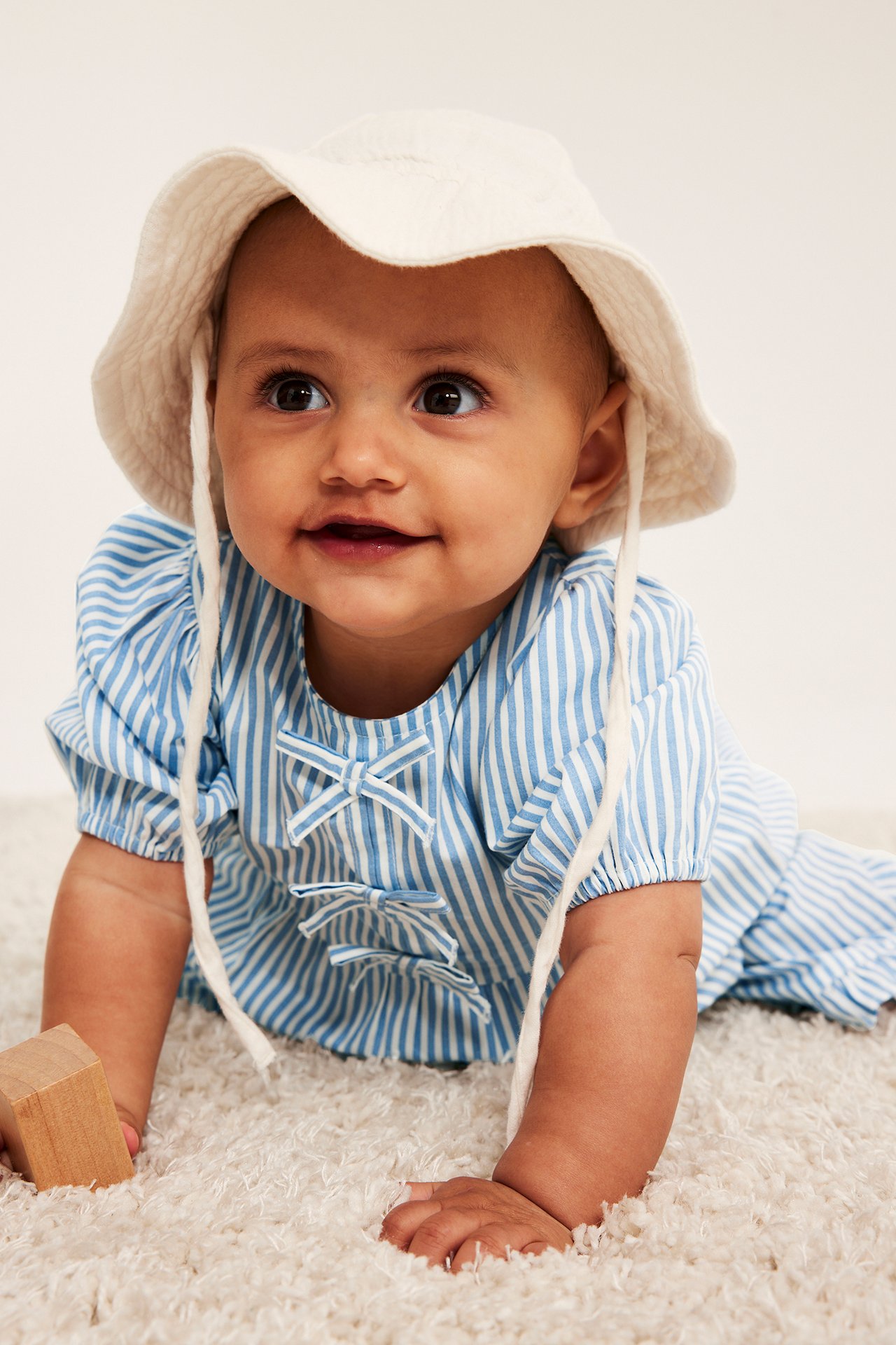 Widok z przodu: niemowlę w jasnoniebieskiej bluzce w paski z bufiastymi rękawami, kokardkami i białym kapeluszu.