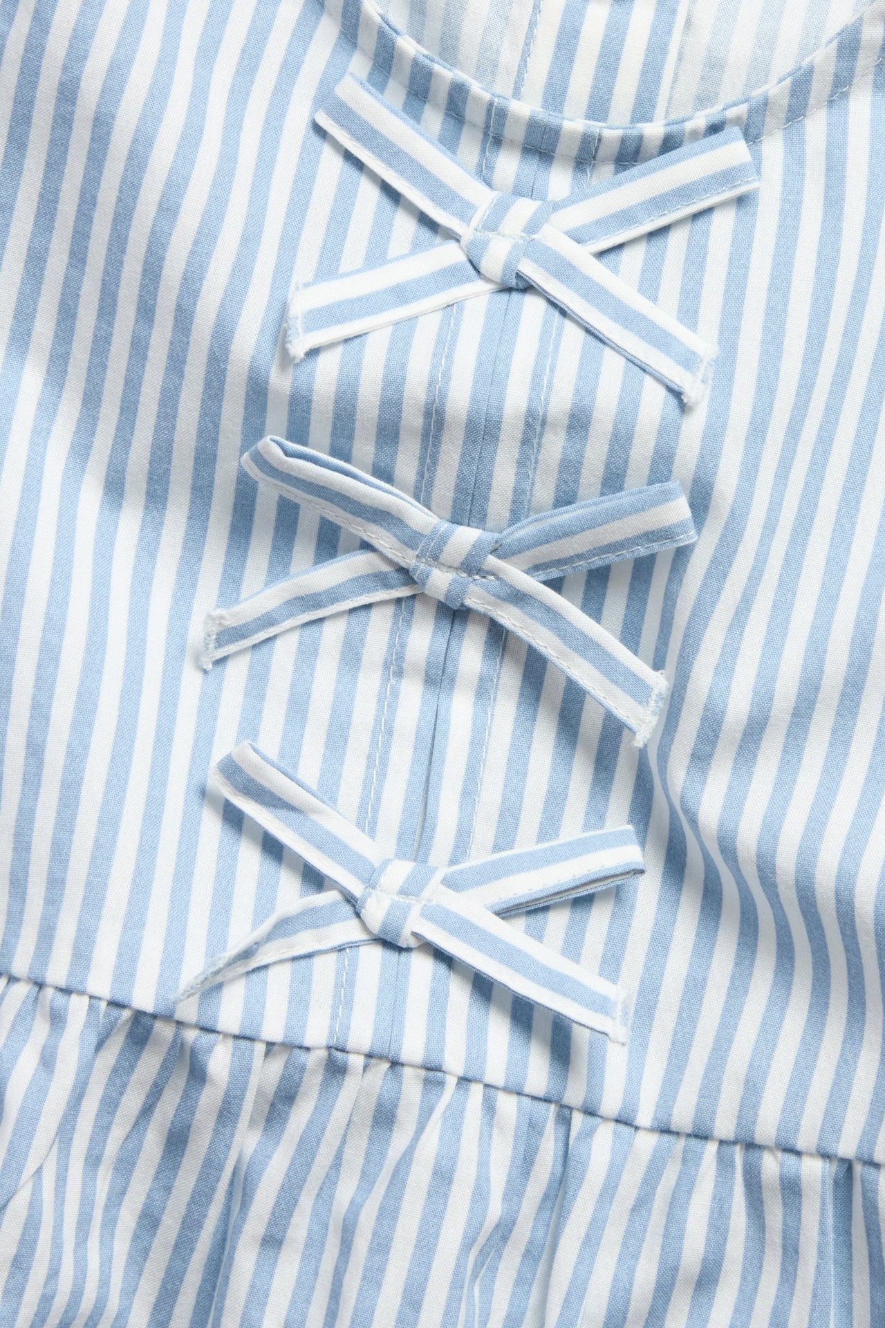 Nærbilde av en babybluse i lyseblå og hvitstripete poplin med tre dekorative sløyfer foran.