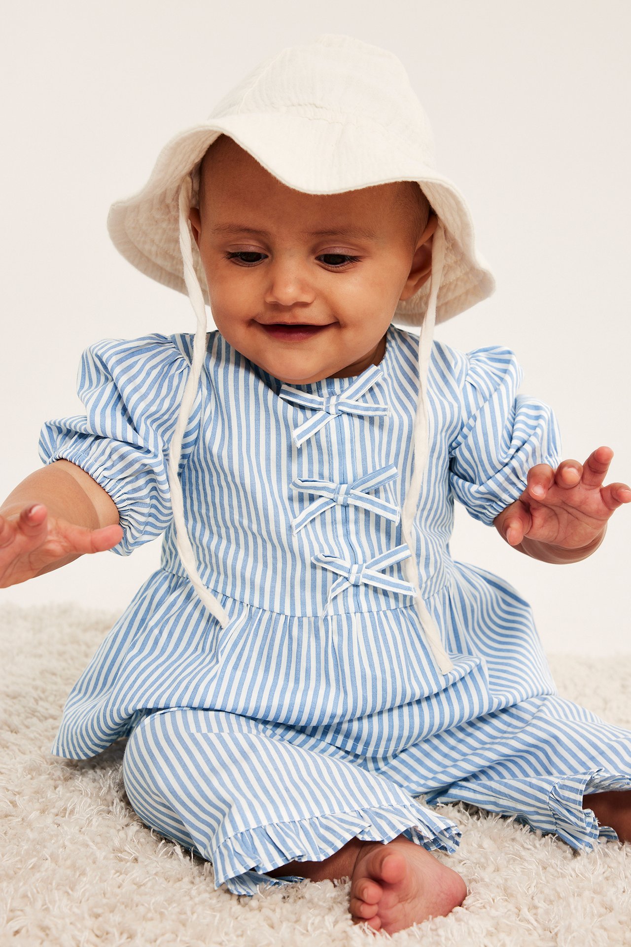 Vauva vaaleansinisessä raidallisessa popliinipuserossa, jossa pussihihat, rusetit, yhteensopivat housut ja valkoinen hattu.