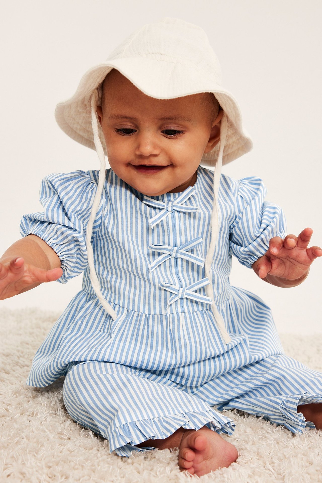 Niemowlę w jasnoniebieskiej bluzce z popeliny w paski, z bufiastymi rękawami, kokardkami, pasującymi spodniami i białym kapeluszem.
