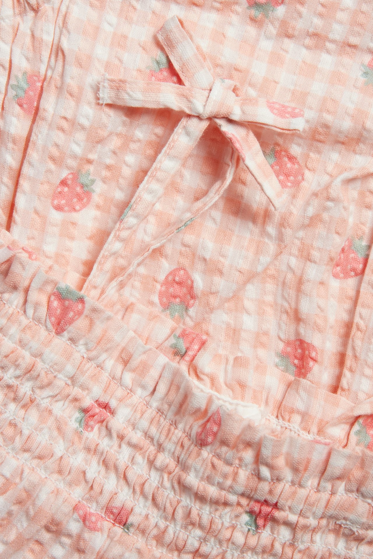 Närbild: Ljusrosa babybody i seersucker med jordgubbsmönster, smock och rosettband.