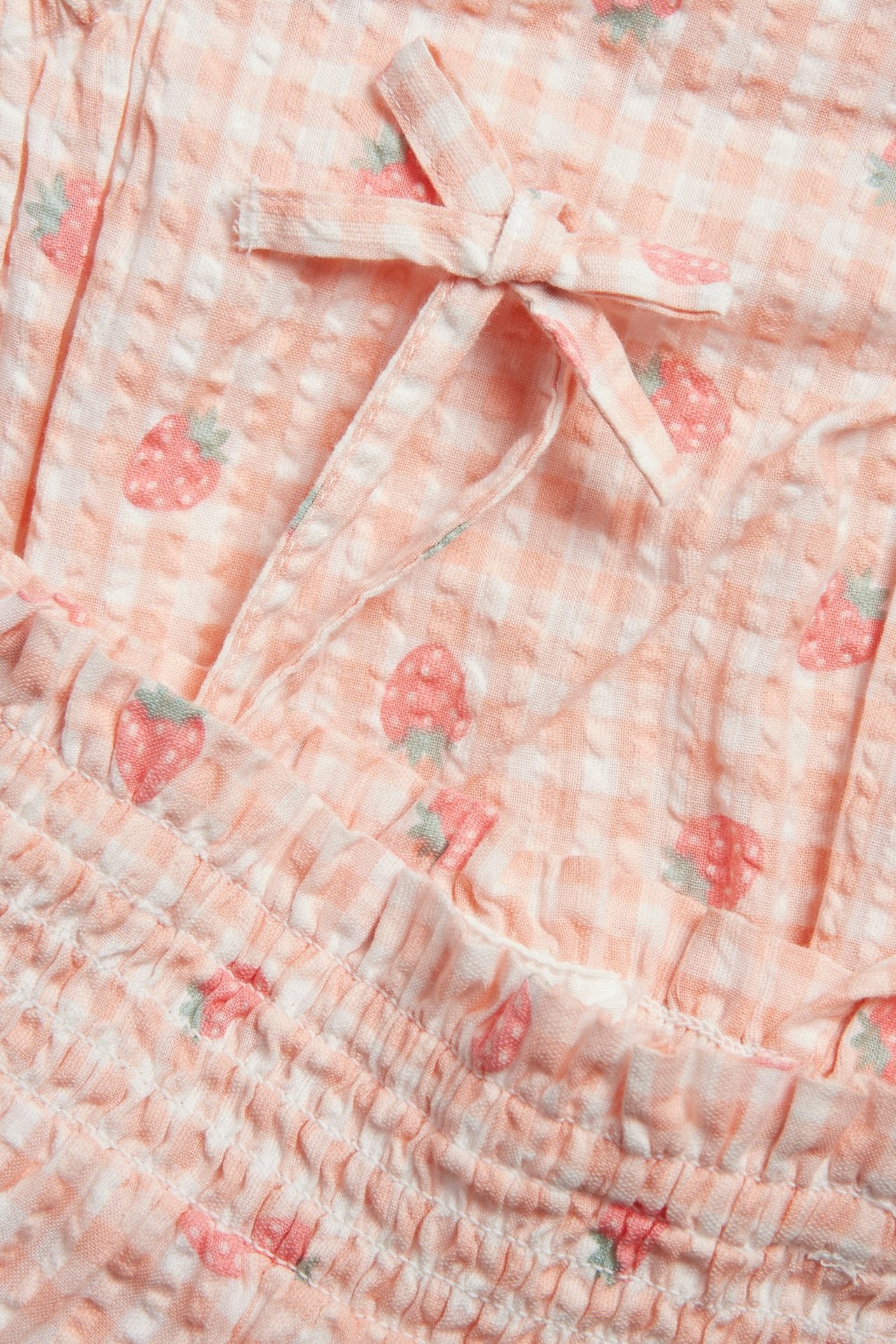 Zbliżenie: Jasnoróżowe body niemowlęce z seersuckeru w truskawki z wzorem gingham, marszczeniami i kokardą.