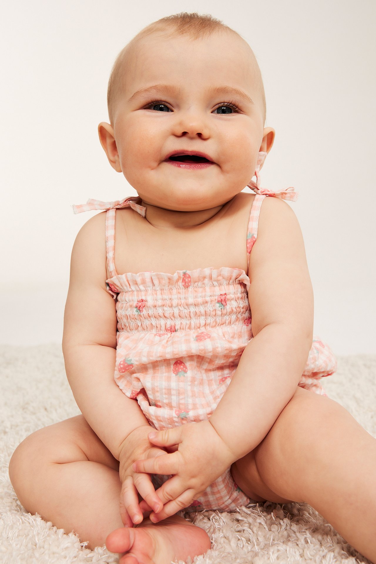 Uśmiechnięte dziecko w jasnoróżowym body seersucker w truskawki z wiązanymi ramiączkami, przód.
