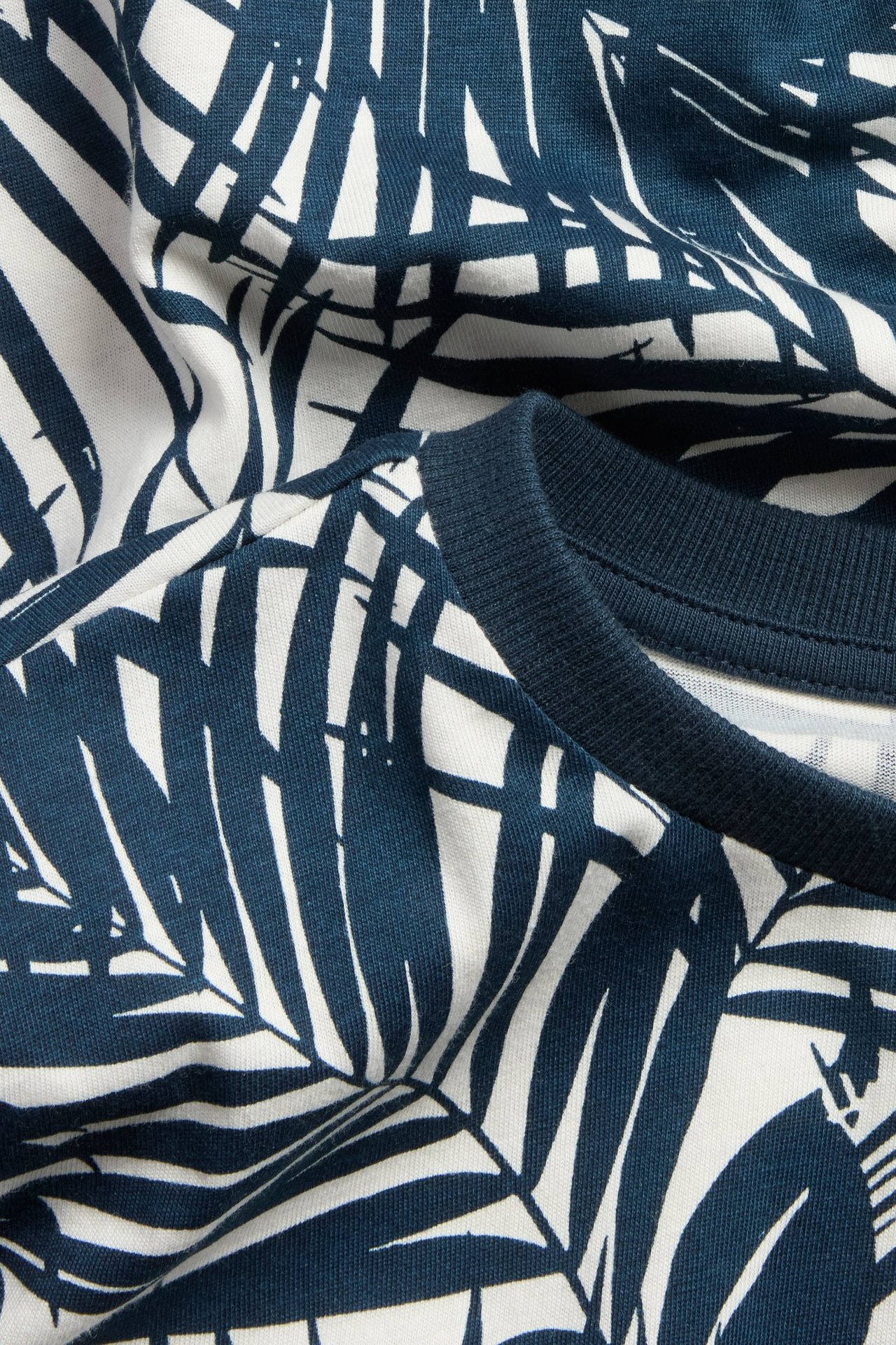 Zbliżenie na bawełniany T-shirt w kolorze złamanej bieli z ciemnoniebieskim wzorem liści i prążkowanym kołnierzykiem.
