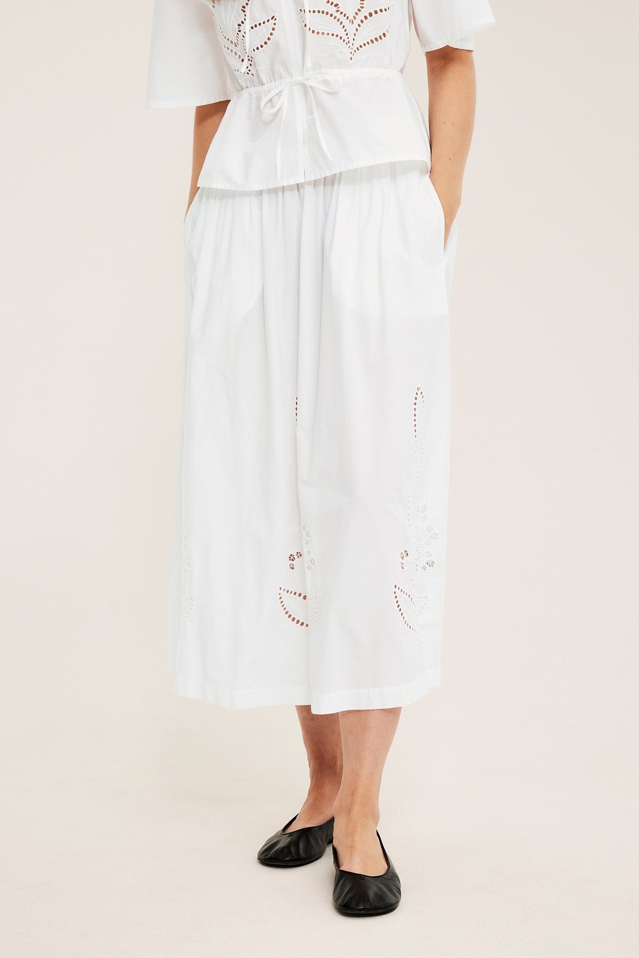 Framifrån: kvinna i vid vit bomullspoplinkjol med brodyr, vit topp och svarta ballerinaskor.