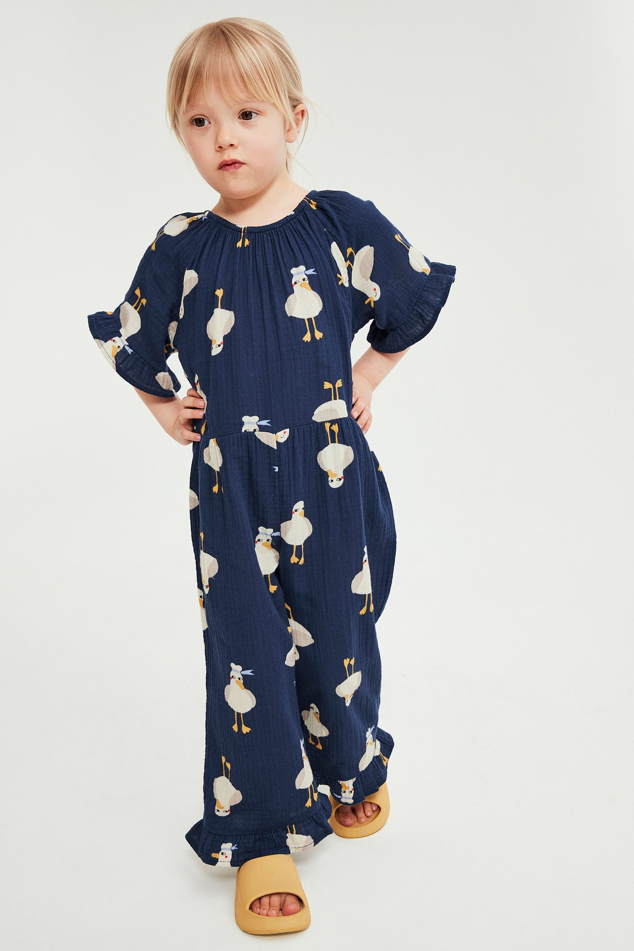 Widok z przodu dziecka w niebieskim, bawełnianym kombinezonie w kaczki i beżowych sandałach.