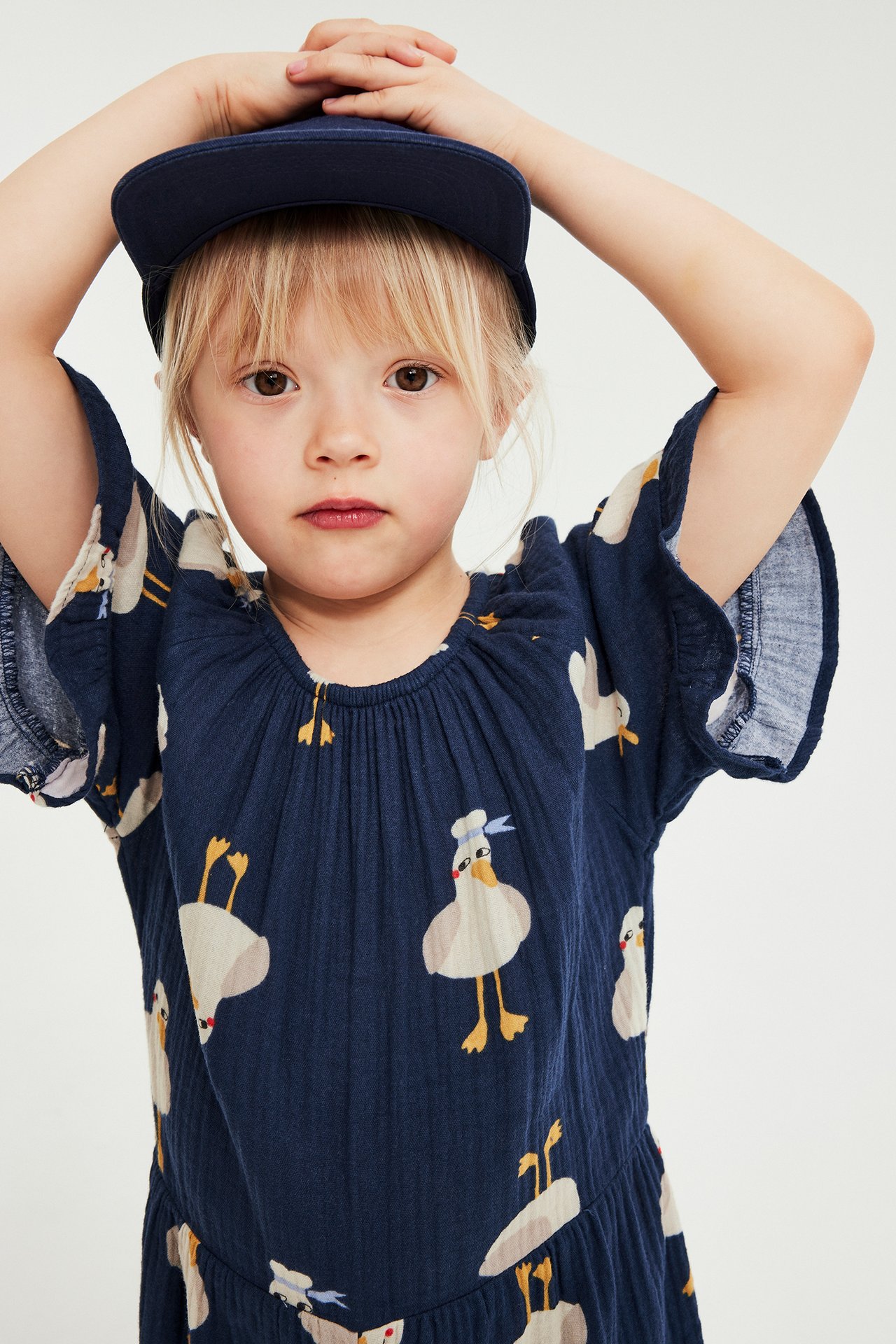 Widok z przodu: Dziecko w niebieskim kombinezonie w kaczki z krótkimi falbankami, nosi granatową czapkę.
