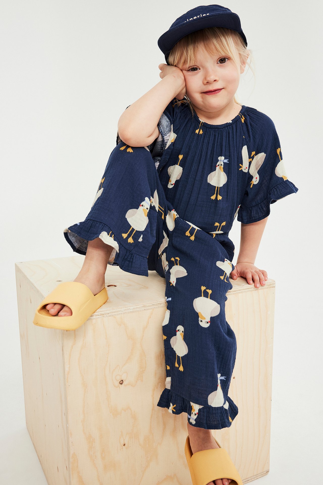 Widok z przodu: Dziecko w granatowym bawełnianym kombinezonie w kaczki, granatowej czapce i żółtych klapkach.