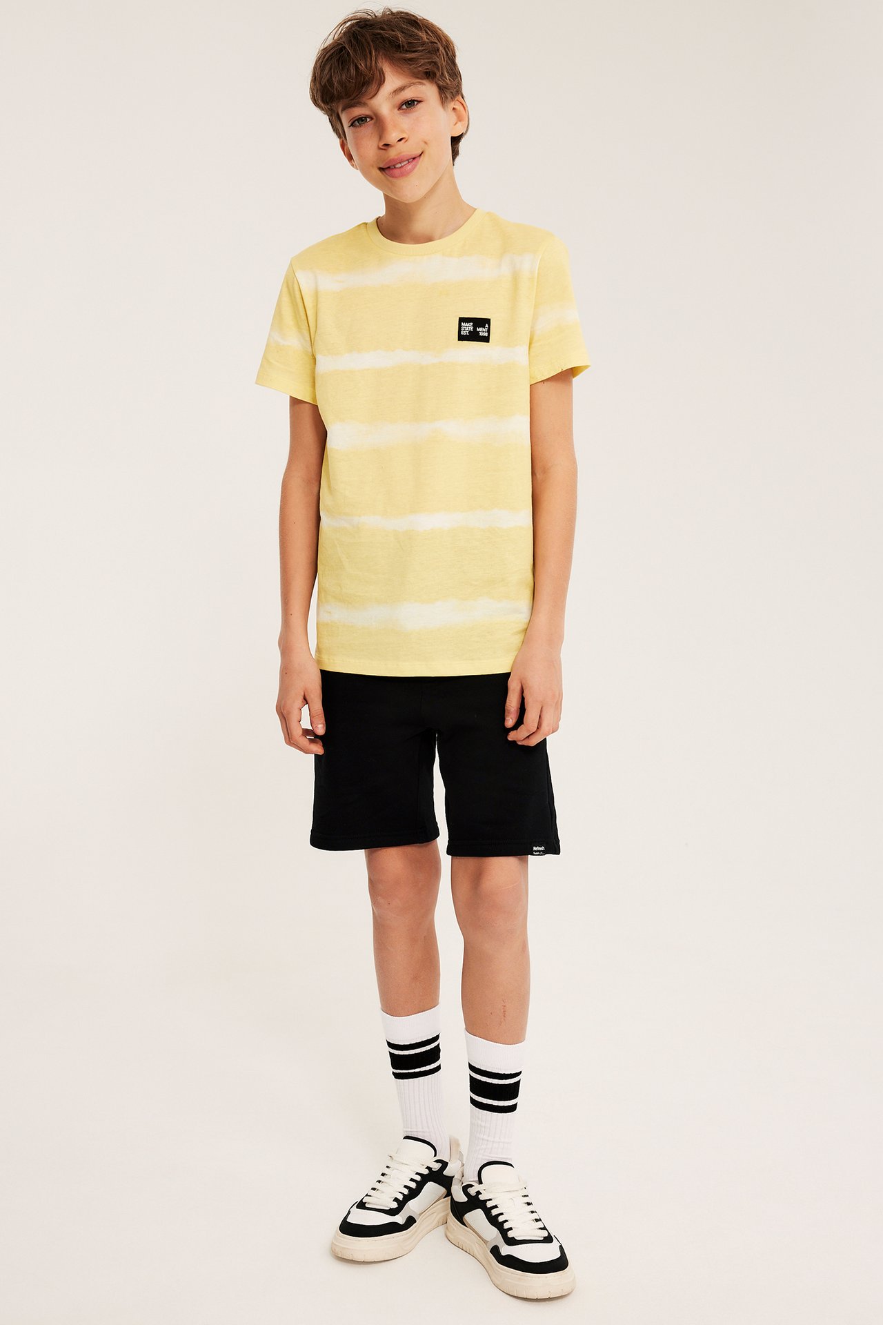 Frontvy: Kid bär en gul batikmönstrad t-shirt, svarta shorts, vita randiga strumpor och vit-svarta sneakers.