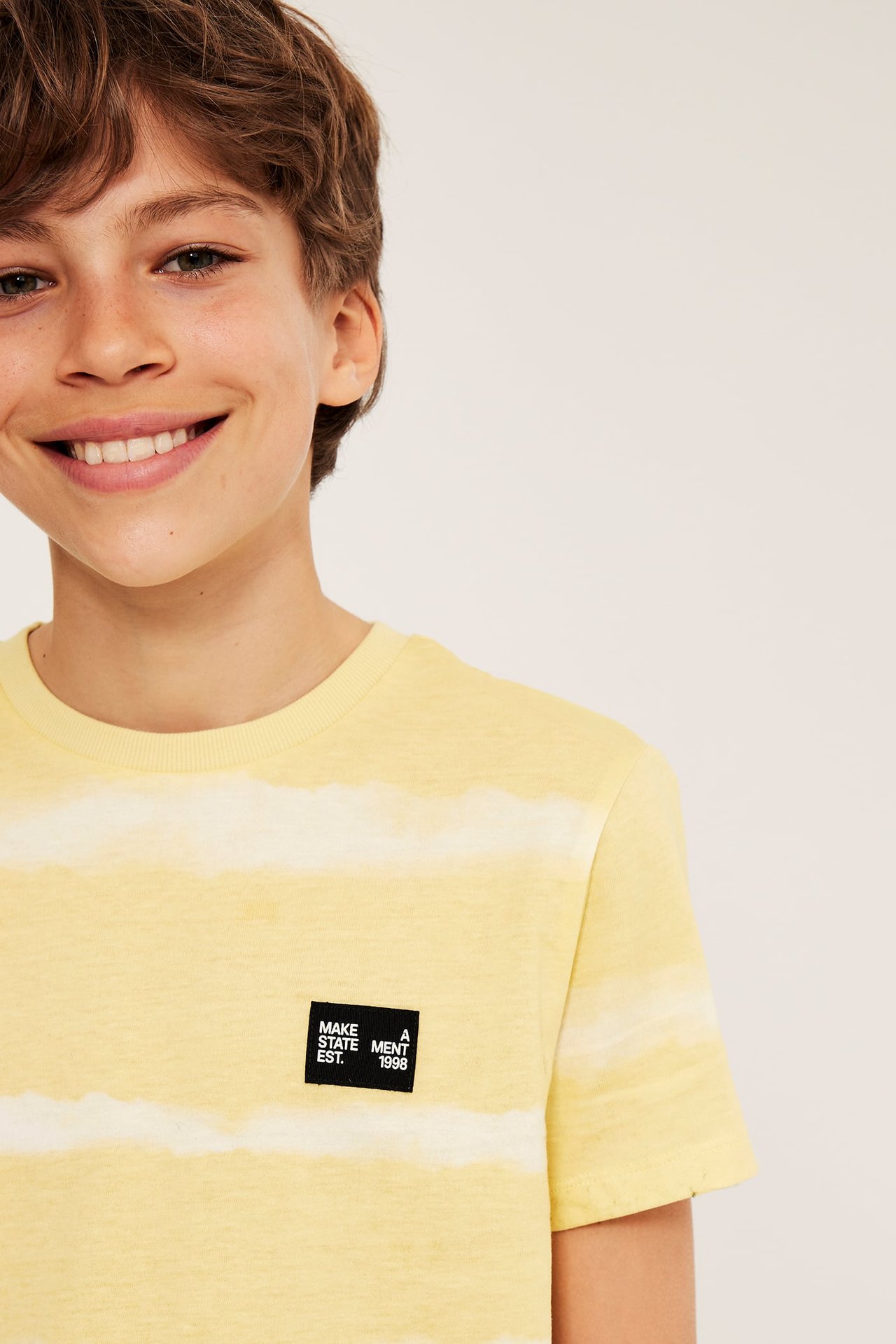 Lähikuva lapsesta, jolla on luonnonvalkoinen ja keltainen batikkikuvioitu puuvilla-T-paita mustalla logomerkillä.