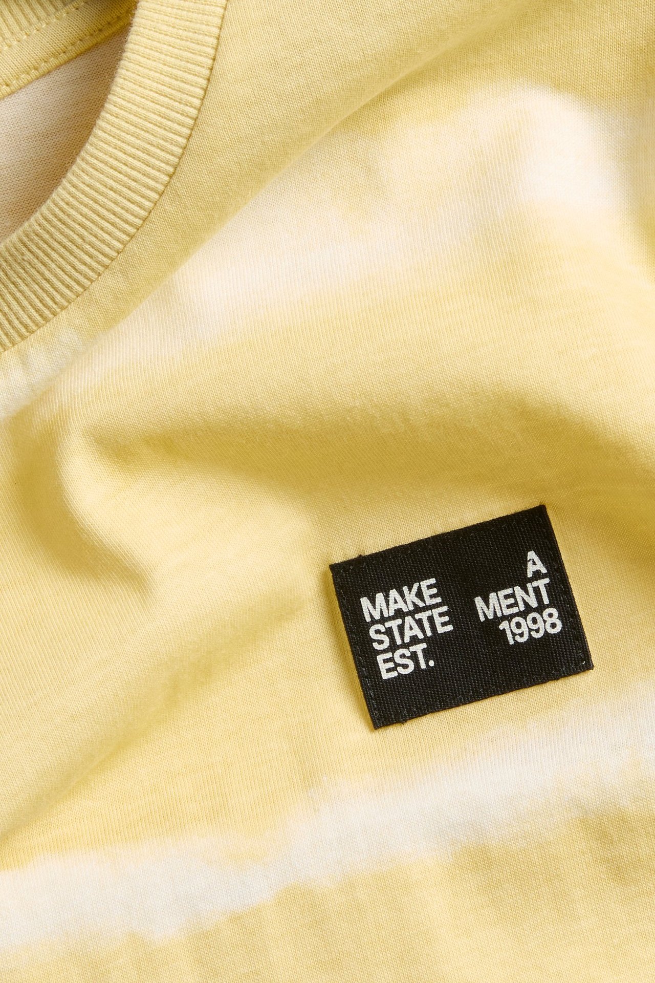 Närbild av en gul och offwhite bomulls-T-shirt med batikmönster och en svart textetikett.