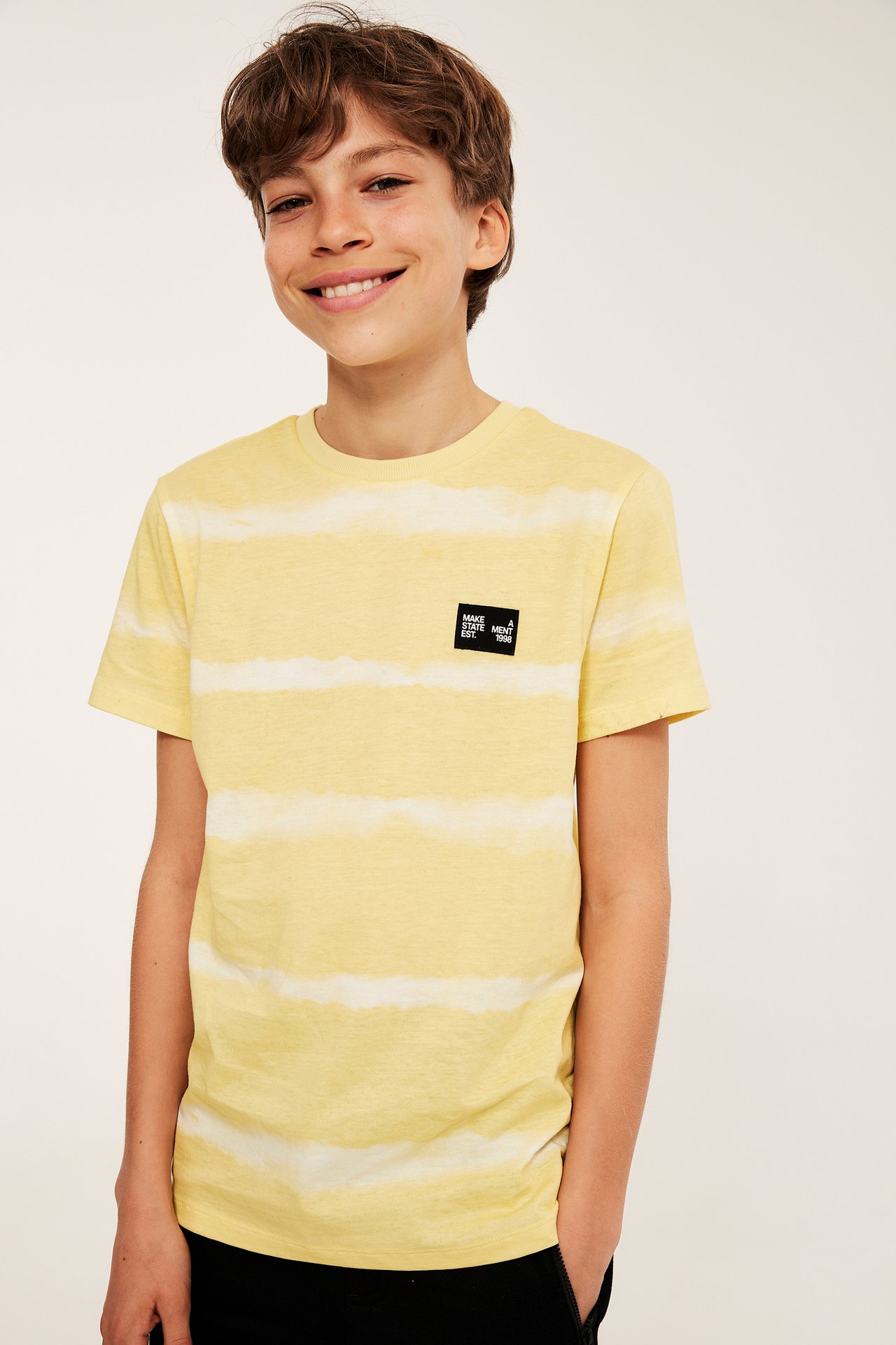 Frontvy av en pojke i en gul och off-white batikmönstrad t-shirt med svart bröstpatch.