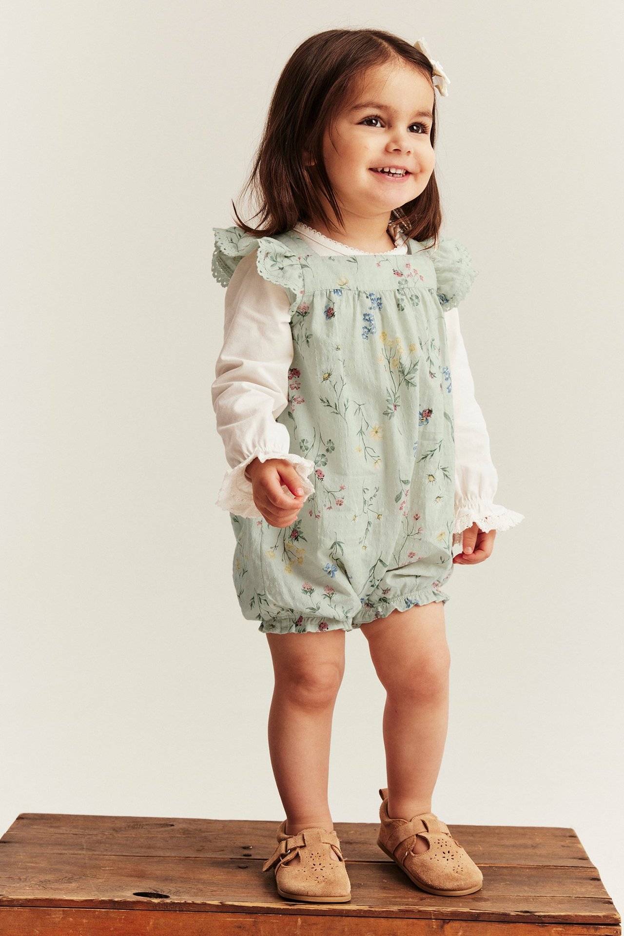 Frontalvisning: Babyjente i lysegrønn blomstret romper, hvit langermet topp og brune semsket sko.