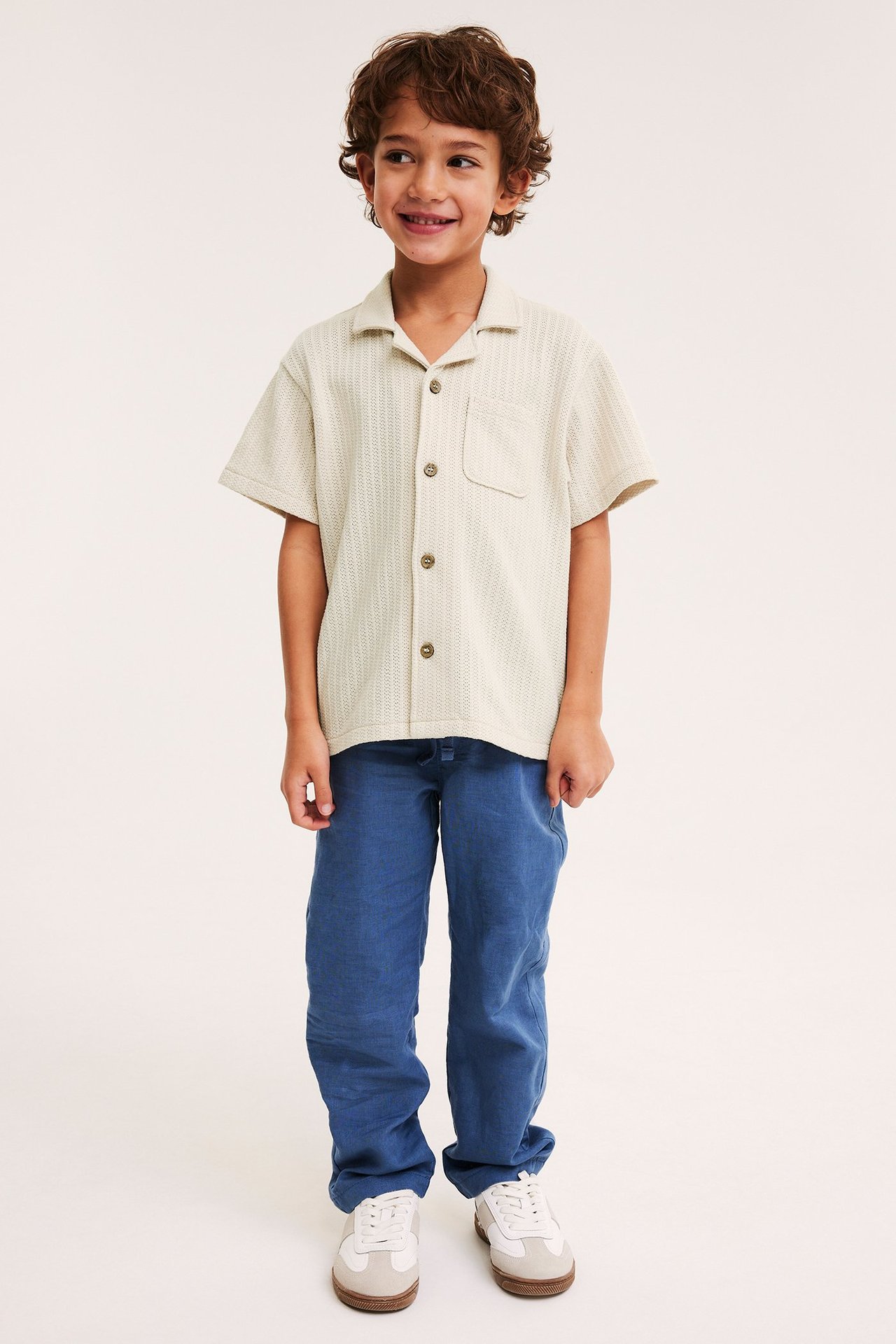 Edestä: Hymyilevä lapsi vaaleabeigessä neulotussa lyhythihaisessa paidassa, sinisissä housuissa ja valkobeigeissä tennareissa.