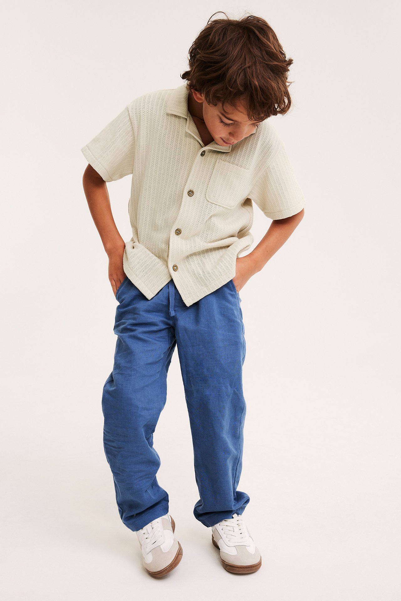Från framsidan: En pojke bär en ljusbeige mönsterstickad kortärmad skjorta, blå byxor och vita sneakers.