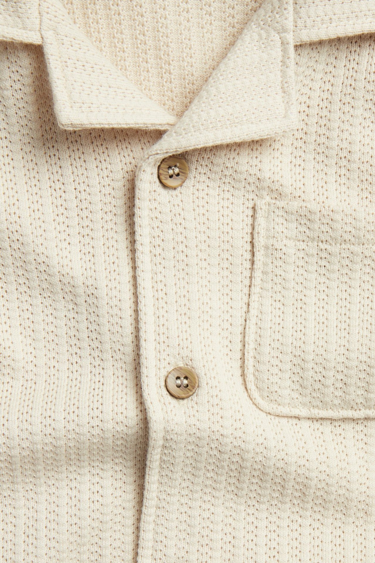Zbliżenie: beżowa dzianinowa koszula z krótkim rękawem, kołnierzem resortowym i drewnianymi guzikami.
