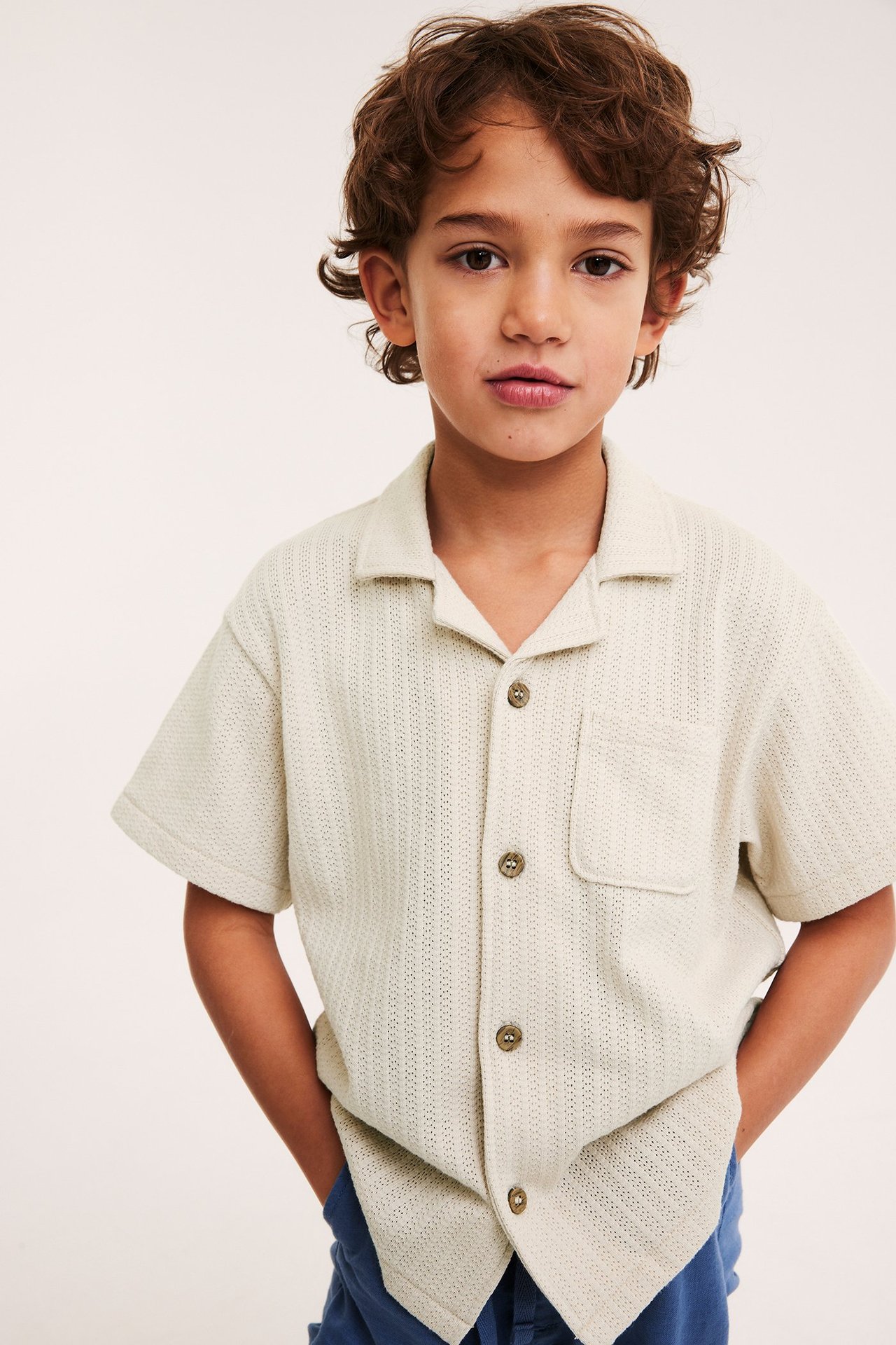 Widok z przodu dziecka w jasnobeżowej dzianinowej koszuli z krótkim rękawem i niebieskich spodniach.