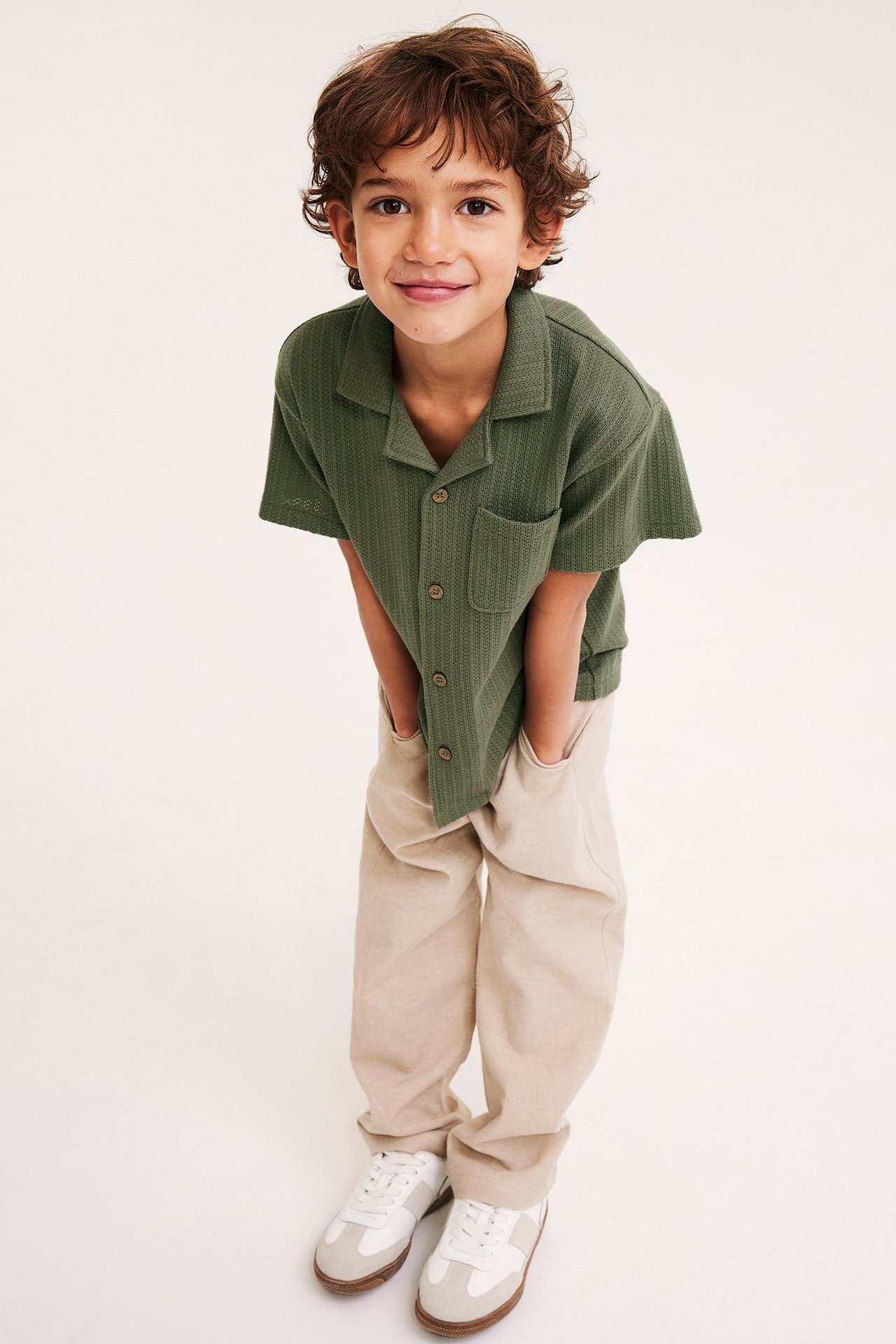 Framifrån, ett barn i en grön kortärmad stickad skjorta, beiga byxor och vita och beiga sneakers.