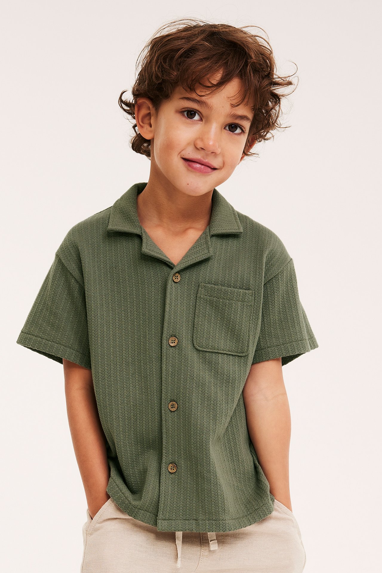 Frontutsikt: barn i grønn strikket kortermet skjorte med resortkrage, treknapper og beige bukser.