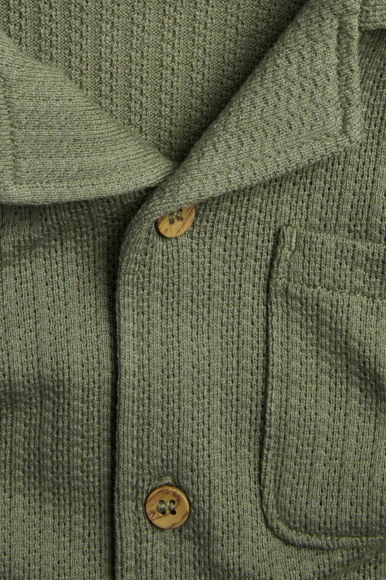 Zbliżenie zielonej dzianinowej koszuli z krótkim rękawem, kołnierzem resortowym, guzikami drewnopodobnymi i kieszenią.