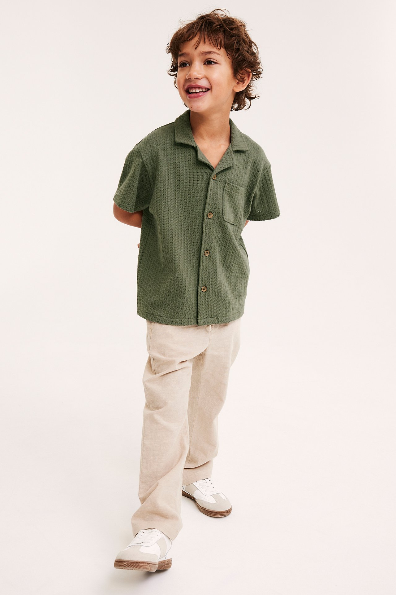 Forfra: Et barn har på seg en grønn mønsterstrikket skjorte med treknapper, beige bukser og hvite joggesko.