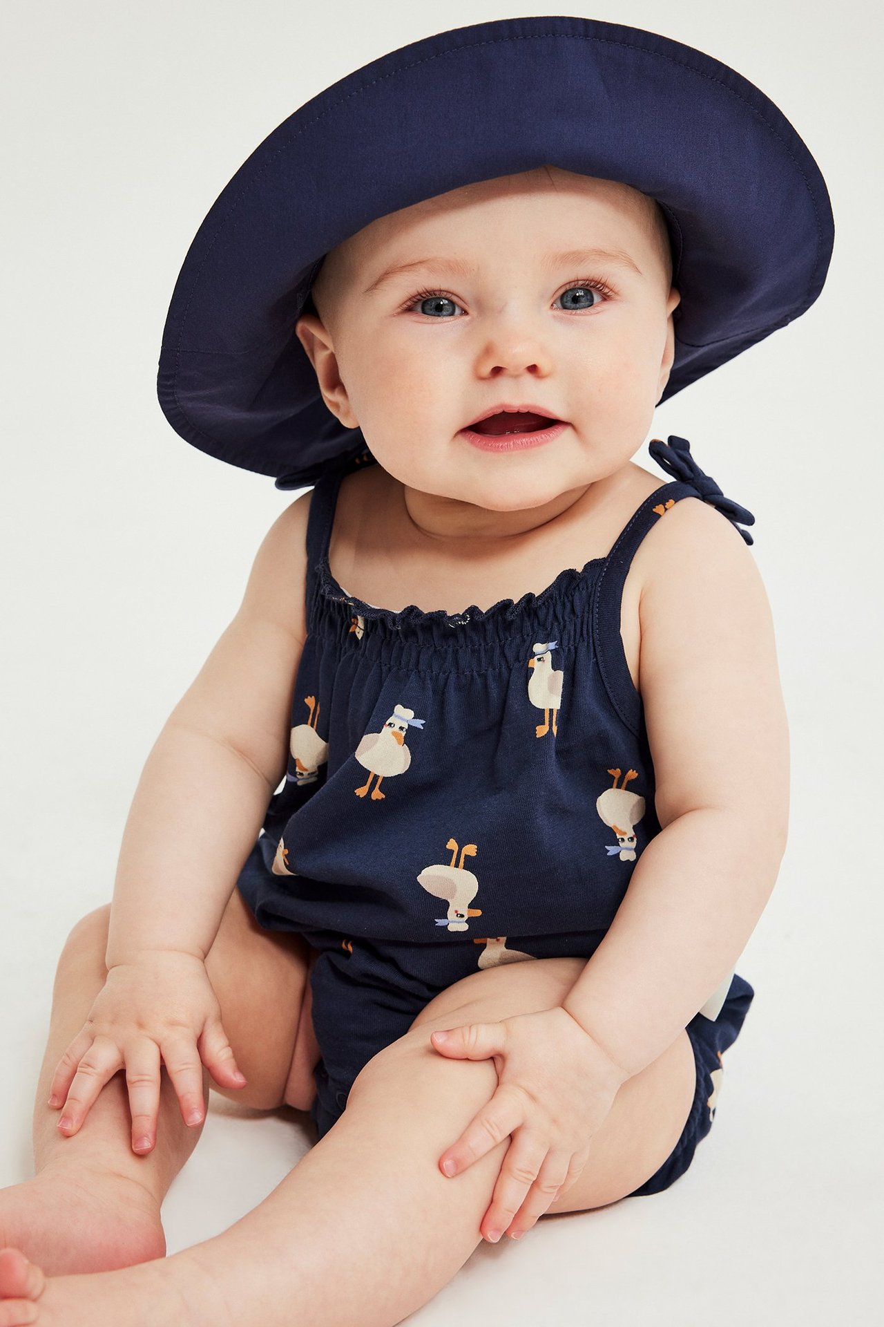 Etunäkymä: vauva tummansinisessä ankka-kuvioisessa puuvillabodyssa rusettinauhoilla ja yhteensopivassa hatussa.