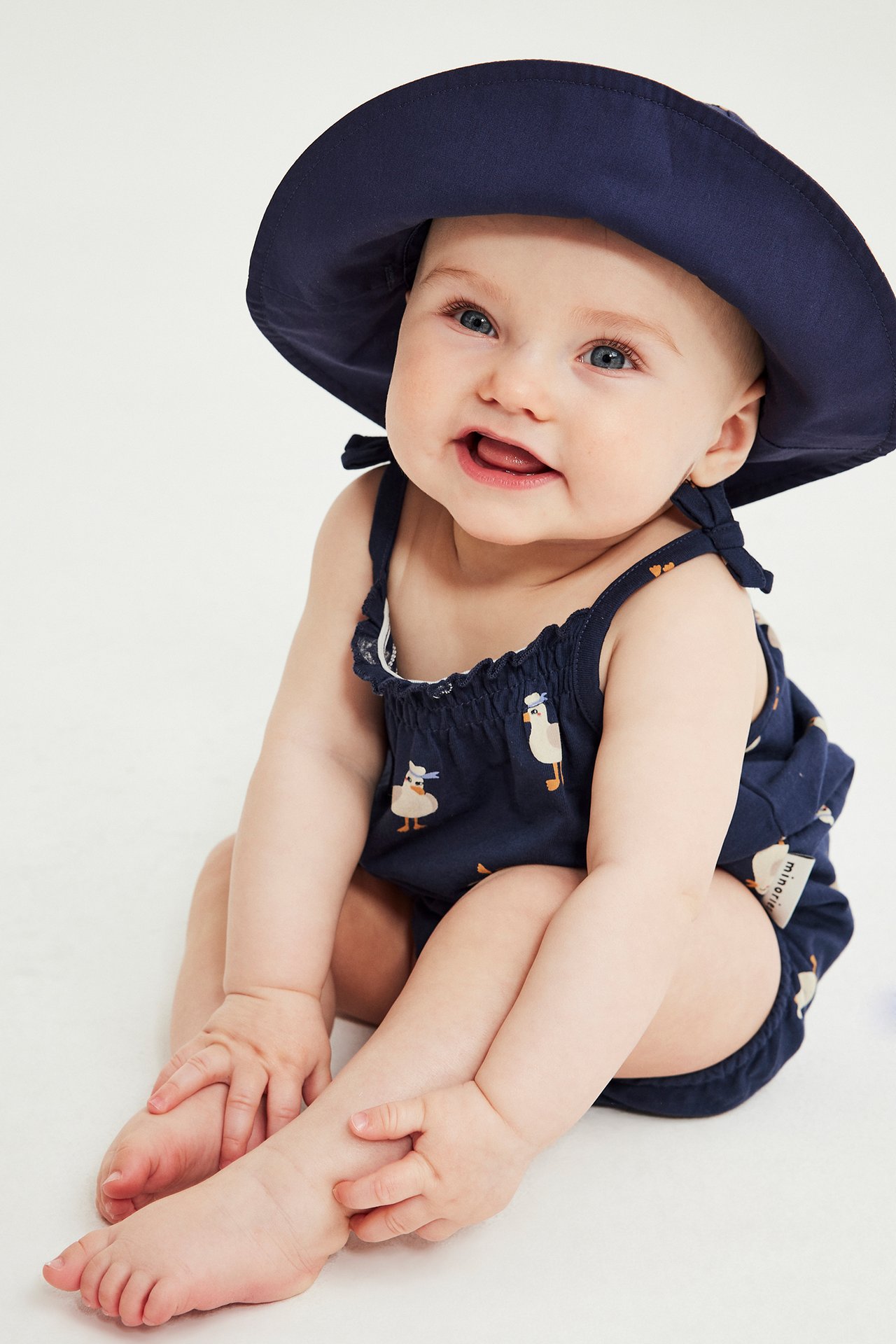 Edestä: vauva yllään tummansininen puuvillainen ankkakuvioinen body ruseteilla ja tummansininen aurinkohattu.